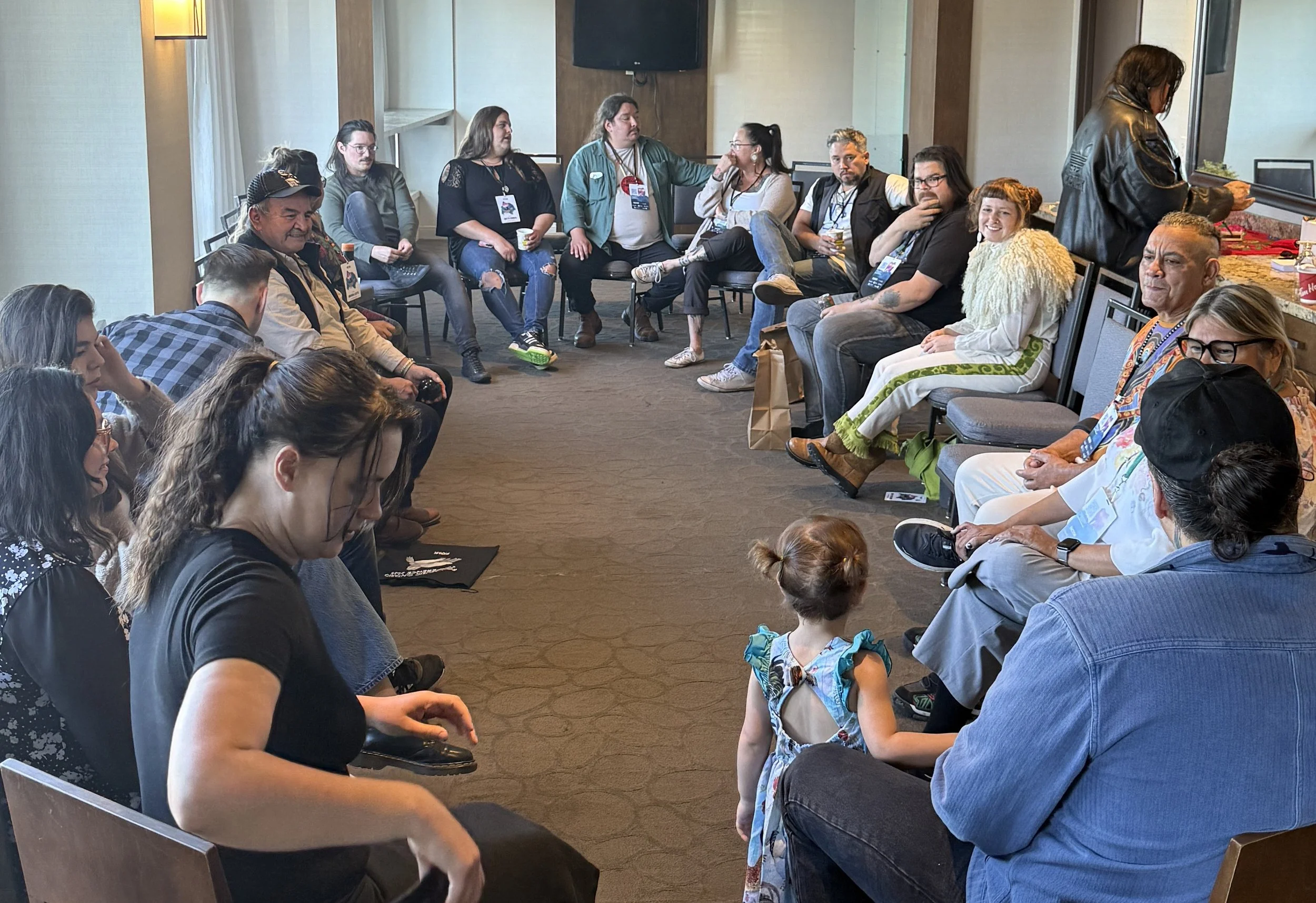 A candid photo of a group of people sitting in a cirlce in a hotel room, casually having a discussion.