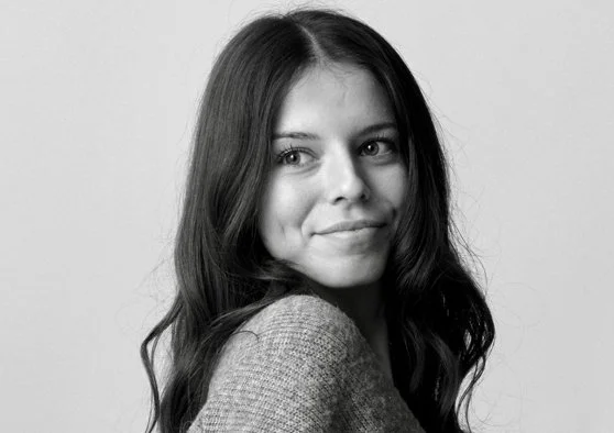 A black & white photo of Taylor Merrick, smiling over her shoulder past the camera, wearing a knitted sweater.