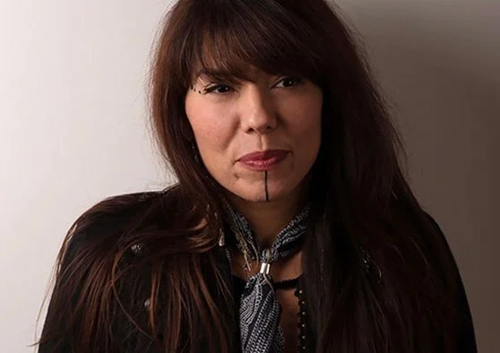 A photo of ShoShona Kish, wearing a cool tie, brown hair, bangs swept to the side. She has a vertical line painted from her lips to her chin and three dots painted on her right temple.