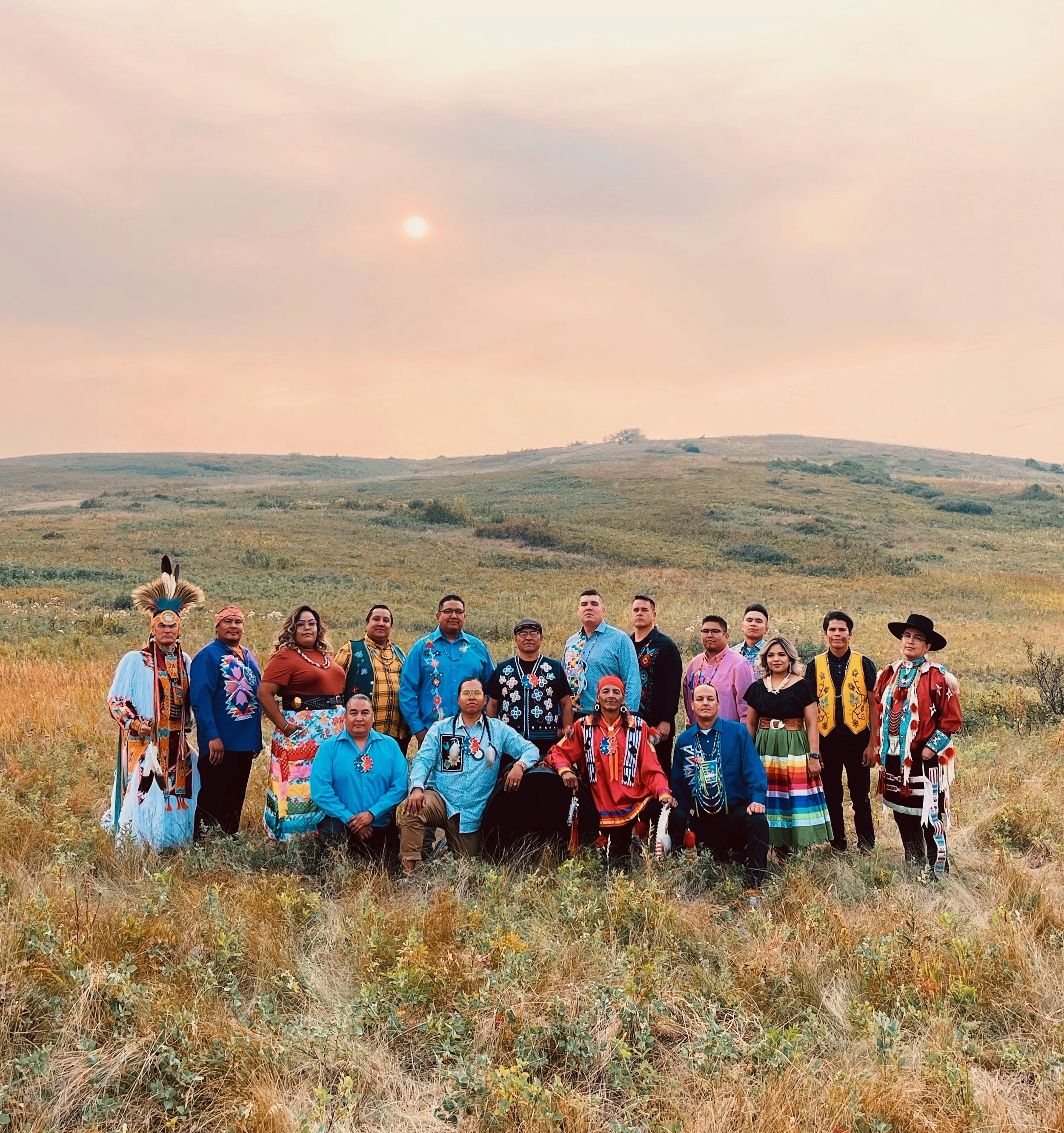 A press photo of Northern Cree, with 16 people posing on grassy plains. They are wearing Indigenous regalia and medallions..