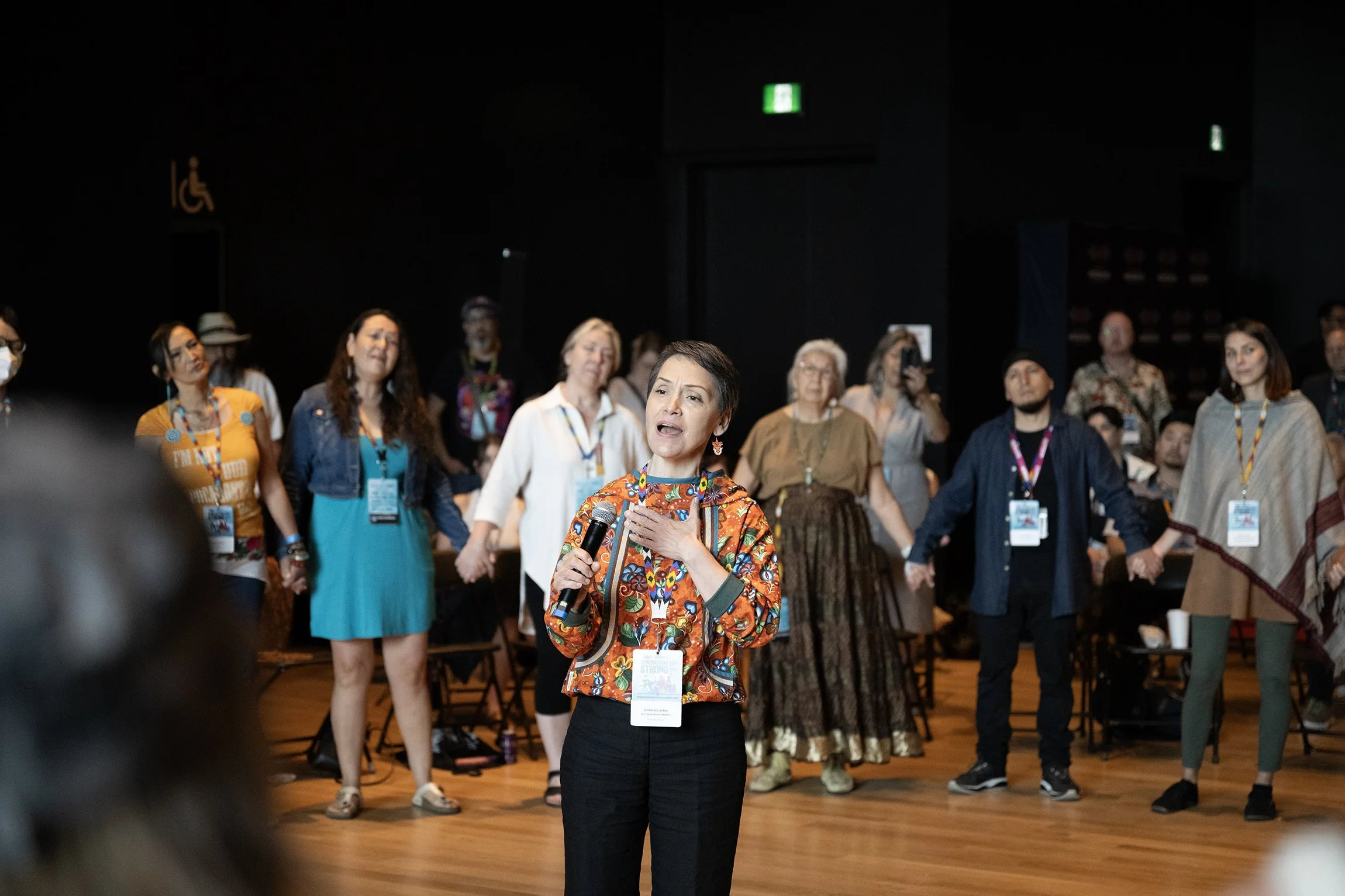 Susan Aglukark is seen singing with a small group of participants around her, holding hands in a circle.