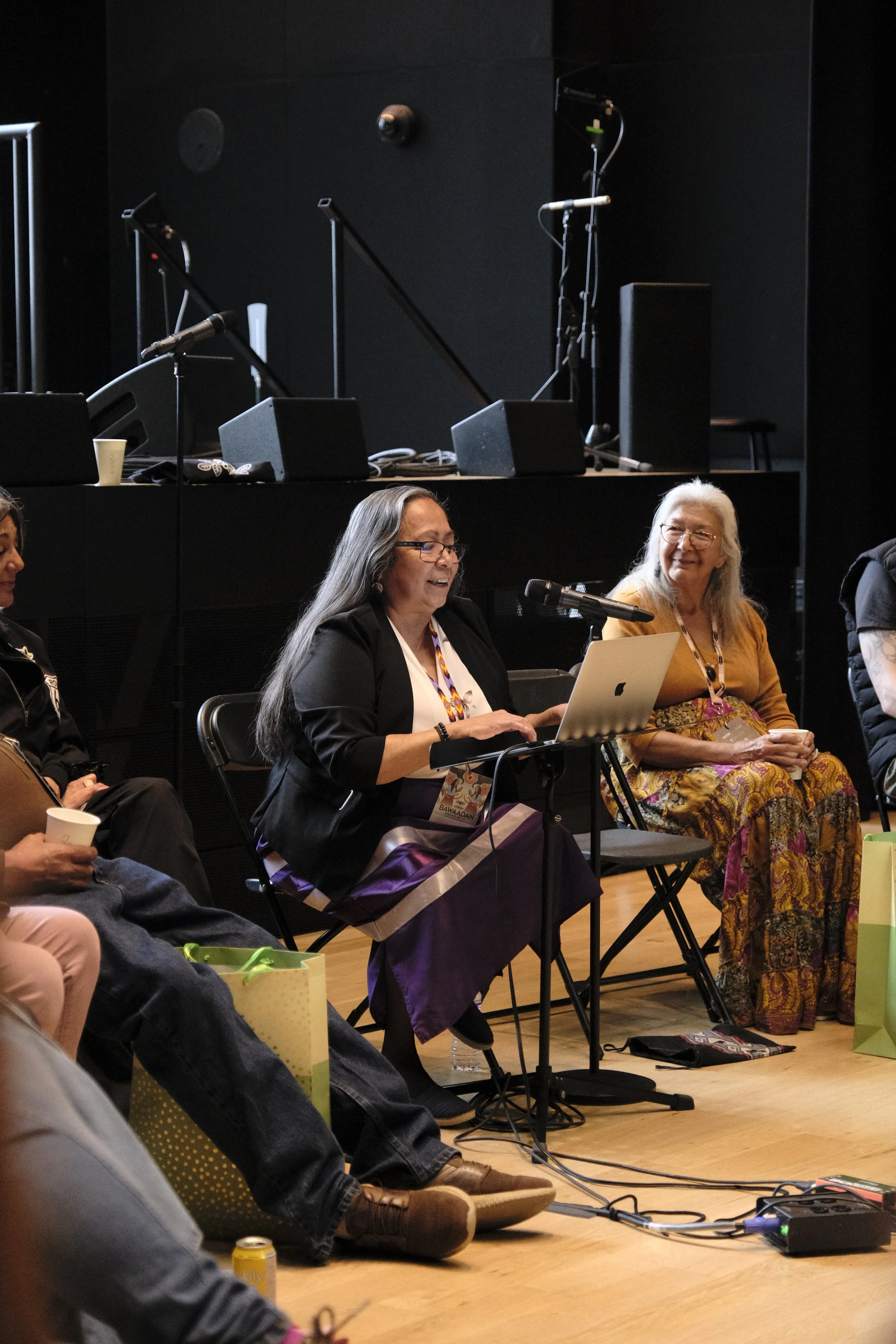 Elder Elaine Bomberry is sitting down, giving a presentation to the crowd during a circle discussion at IIMS. Elder Louise Lahache is sitting beside Elaine, looking at her and smiling affectionately.
