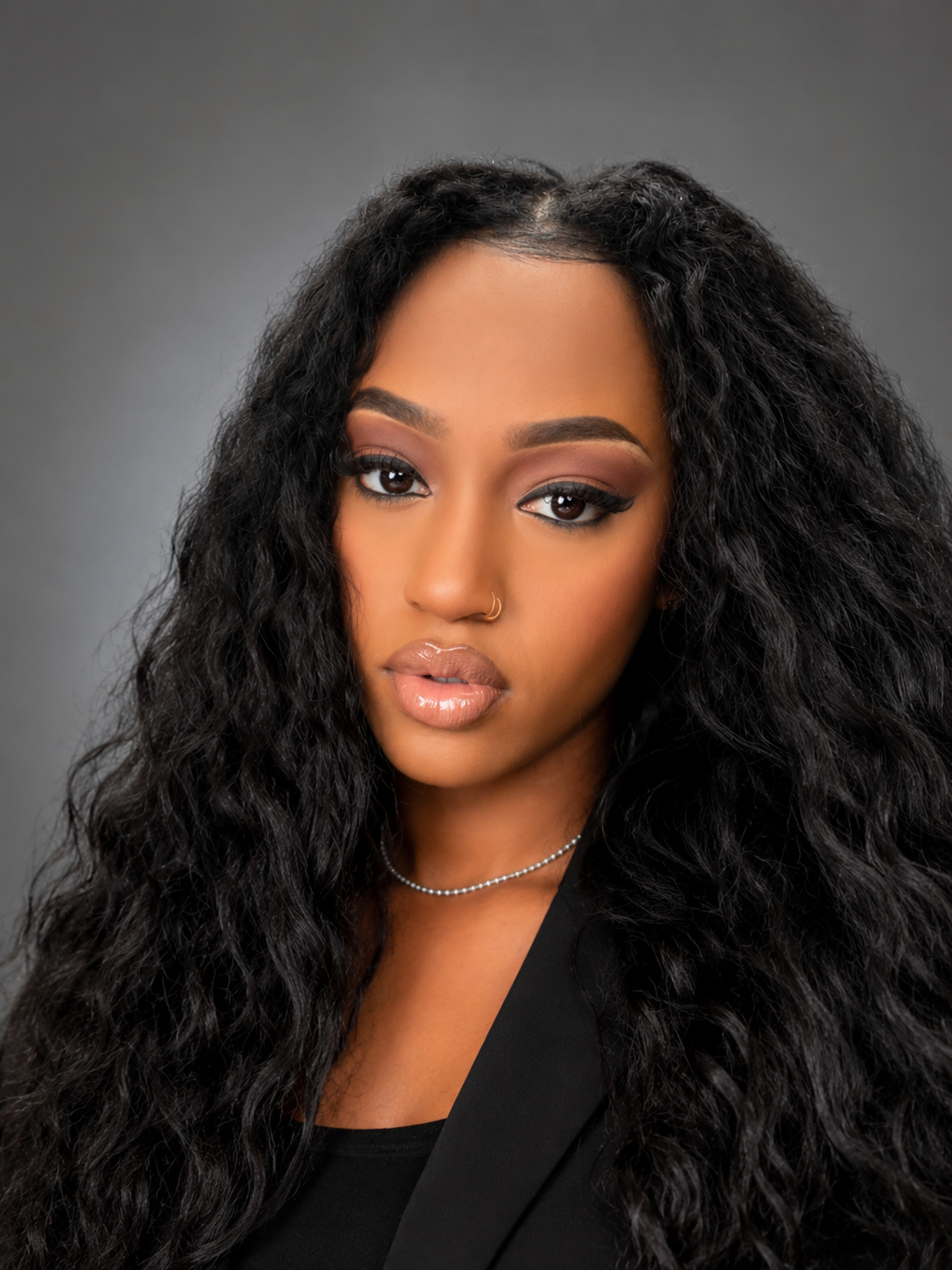 A young woman with long, curly black hair, wearing makeup with nude lips and eye makeup, a small nose ring, a black outfit, and a delicate silver necklace, against a gray background.