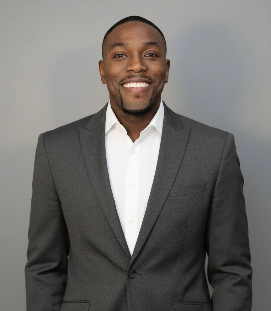 A smiling man in a dark gray suit with a white shirt against a plain gray background.