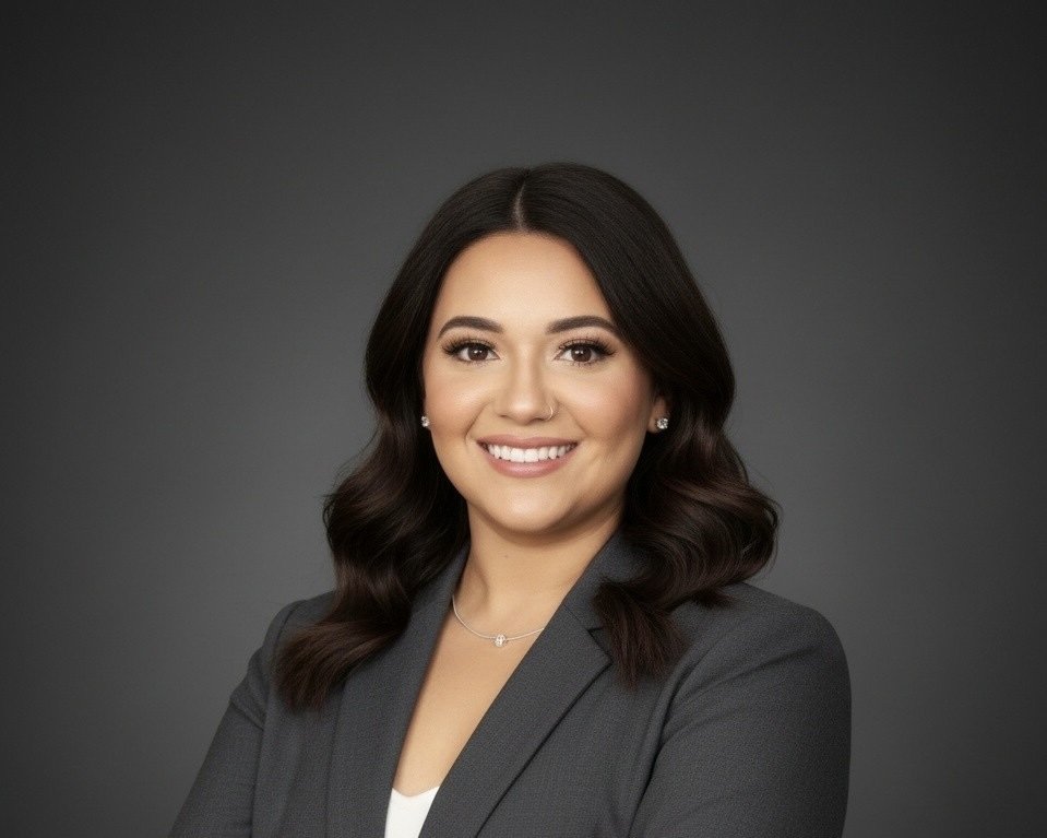Portrait of a smiling woman with dark hair, wearing a gray blazer and jewelry, against a dark gray background.