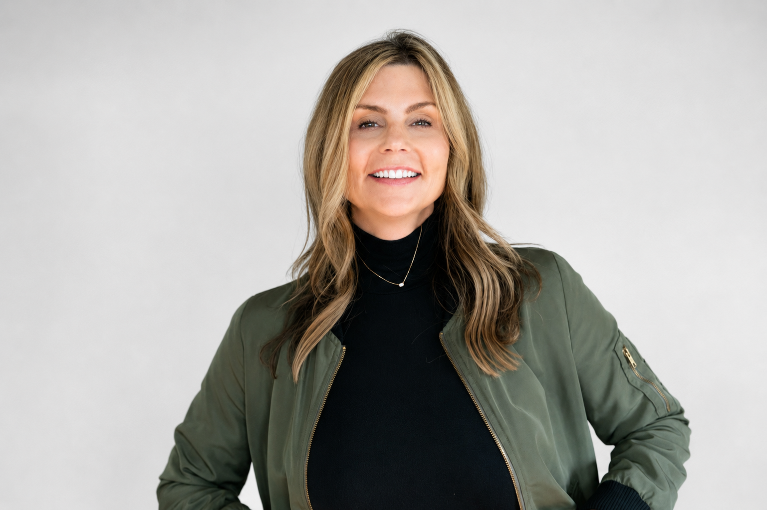 Headshot of a smiling woman with long wavy hair, wearing a black turtleneck, a green bomber jacket, and a delicate necklace, standing against a plain light background.