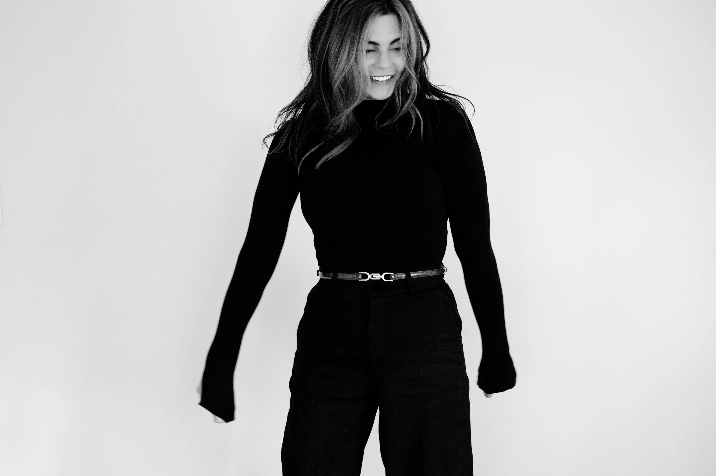 Black-and-white photo of a smiling woman looking down, wearing a black turtleneck and black pants with a belt, against a plain light background.
