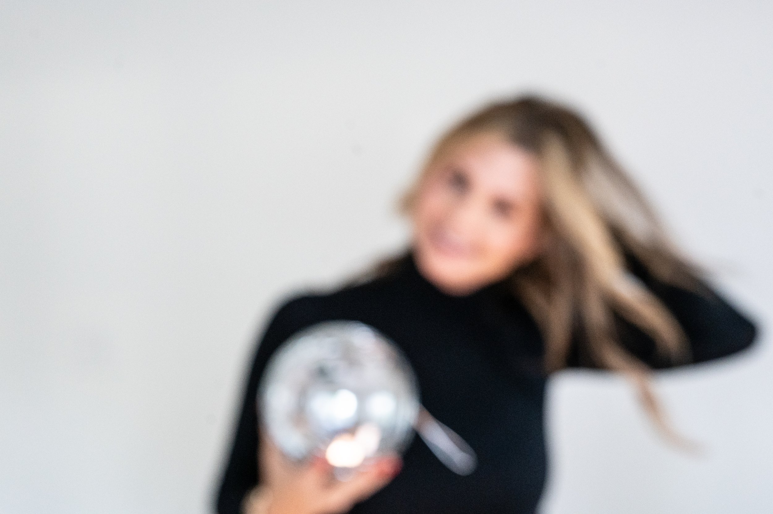 A person with long wavy hair smiling, wearing a black top with white striped sleeves, holding a shiny circular object with colorful markings in front of a plain white wall.