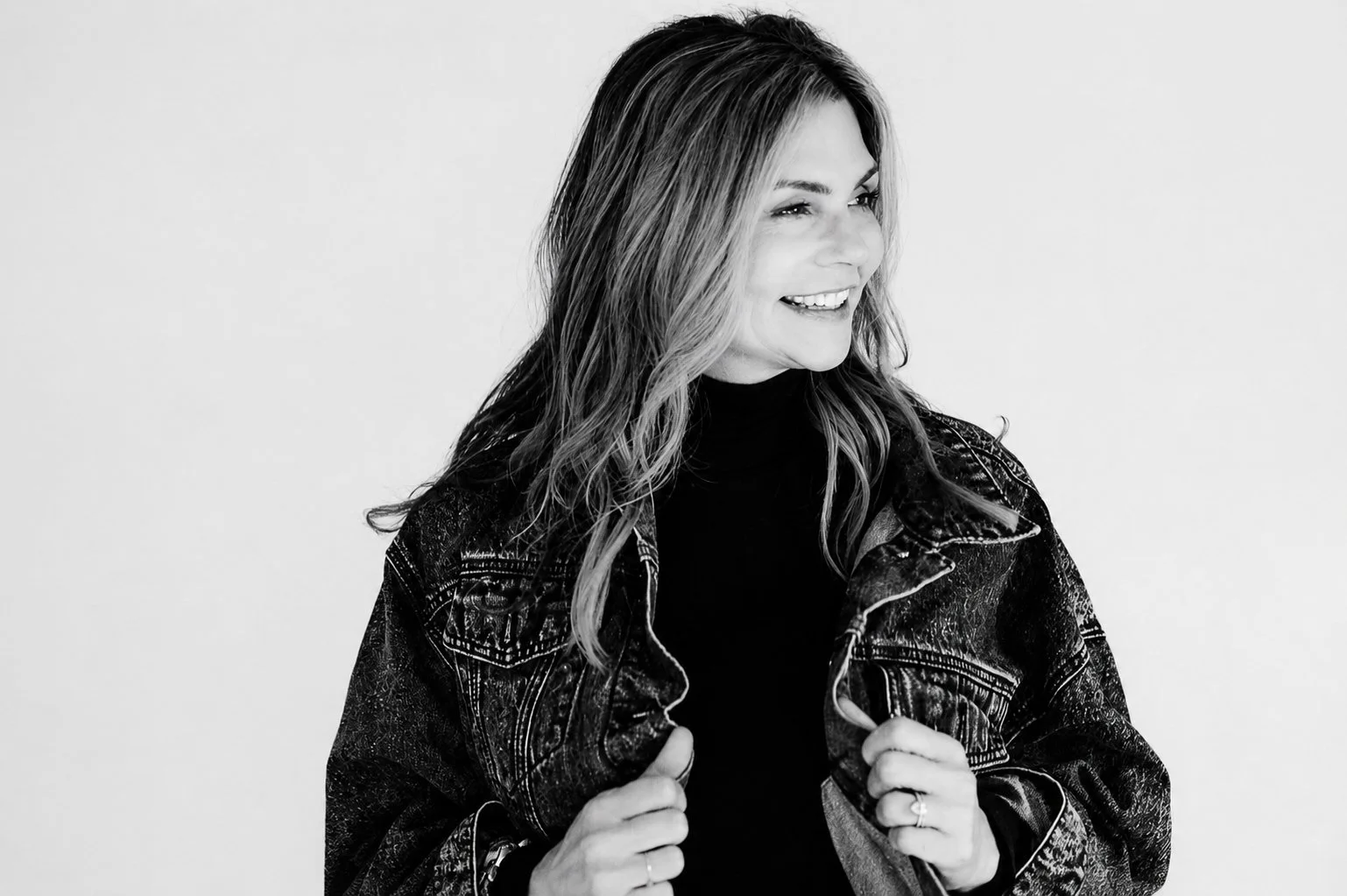 A black-and-white photo of a woman with wavy hair, smiling and looking to her right, dressed in a denim jacket over a black top, holding the lapels of her jacket.