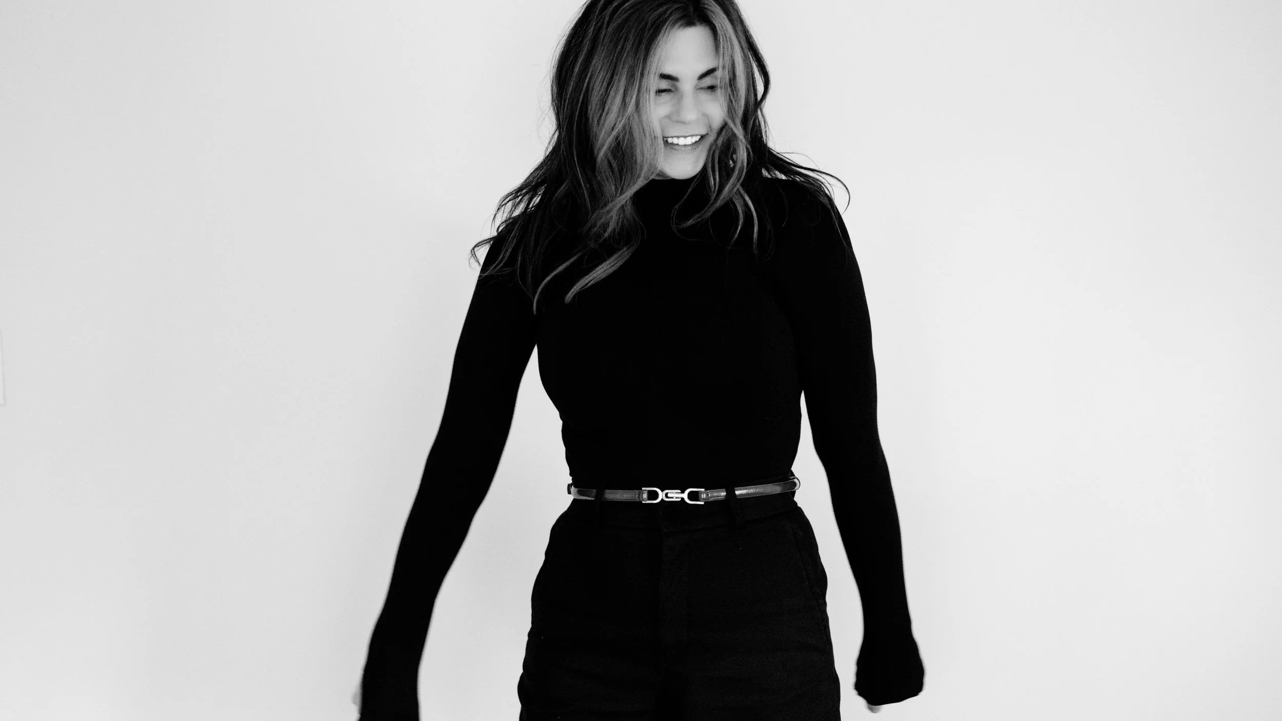 A black-and-white photo of a woman with long hair, smiling and looking down, wearing a black turtleneck and a belt.