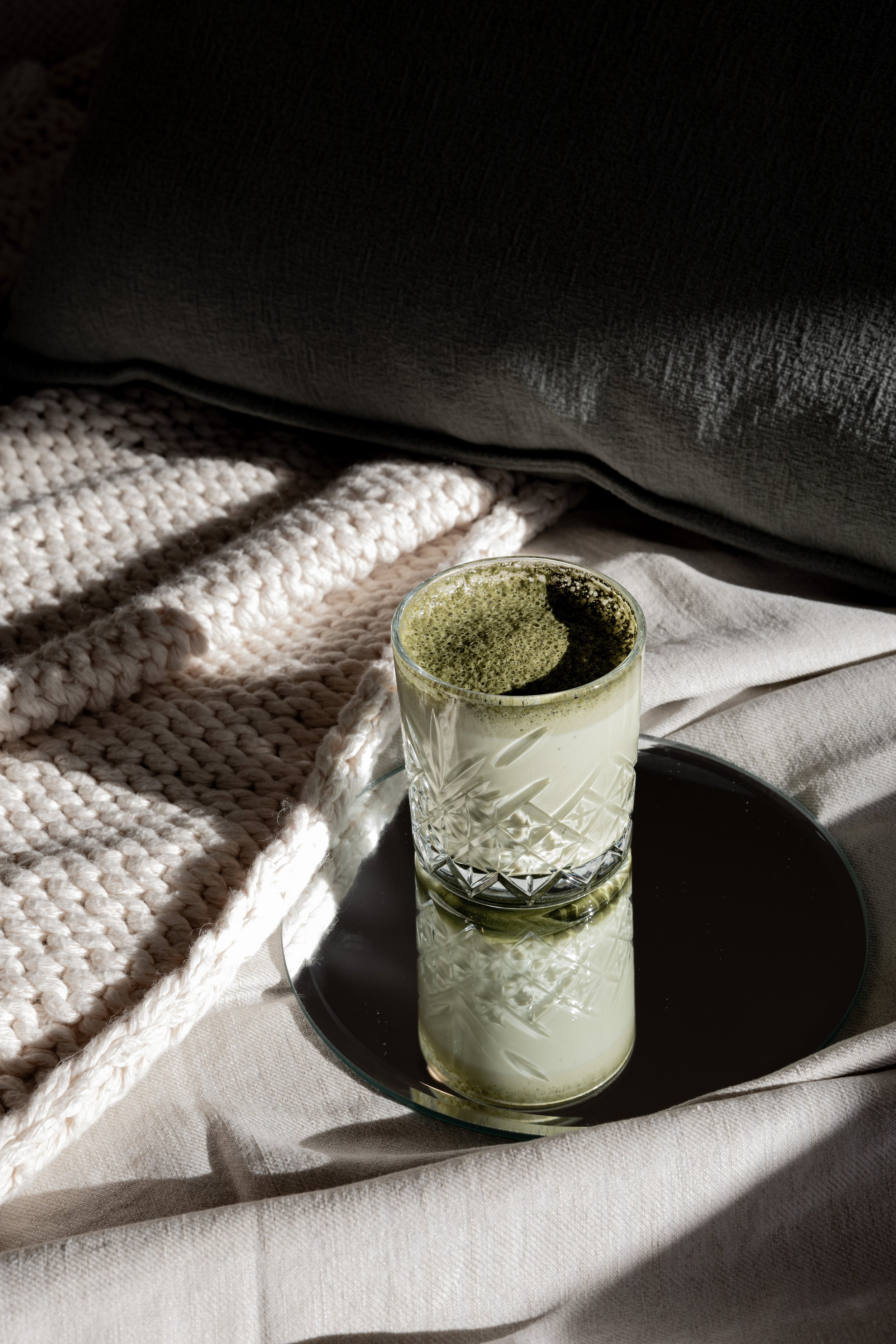 Glass of matcha latte on a mirrored tray, placed on a bed with beige and gray bedding, illuminated by natural sunlight.