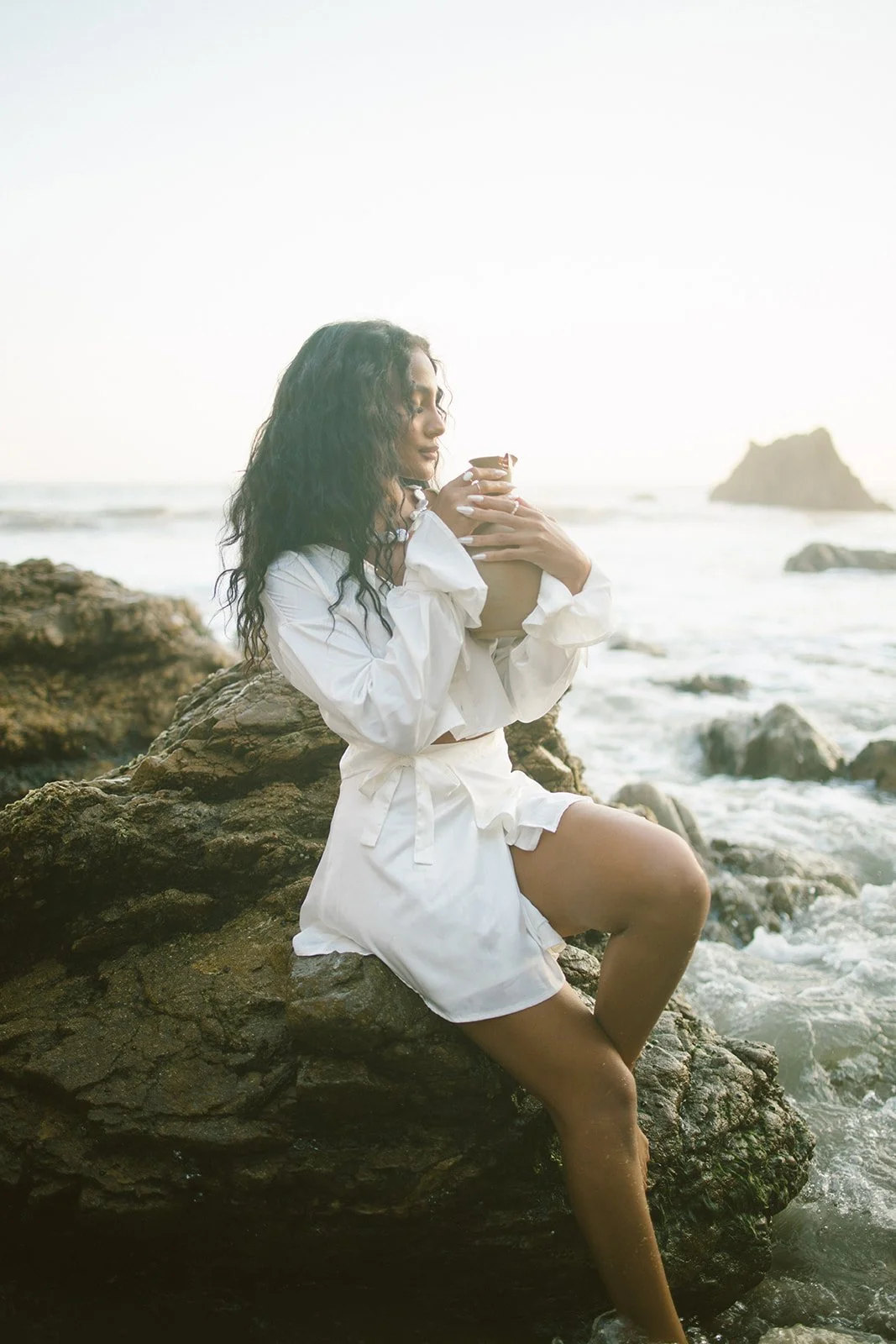 Woman sitting on a rock by the ocean, holding a cup, wearing a white outfit, with waves and rocks in the background.
