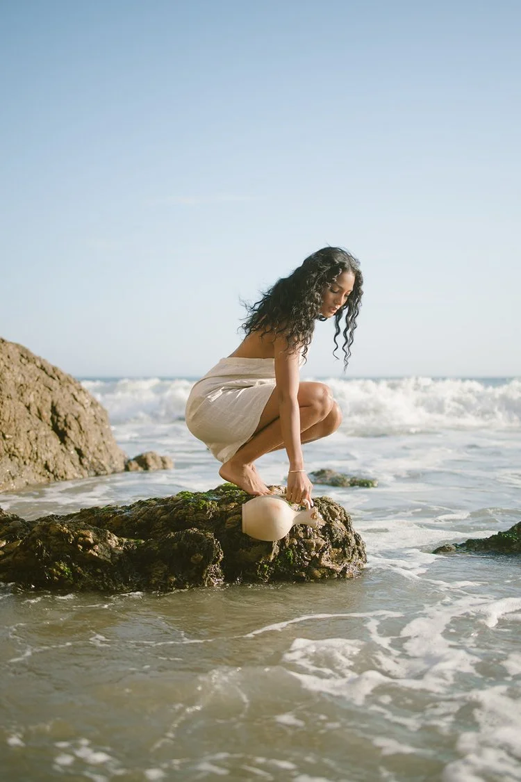 Woman with curly hair crouching on a rock at the beach, holding a white ceramic teapot, with waves in the background and a clear blue sky.