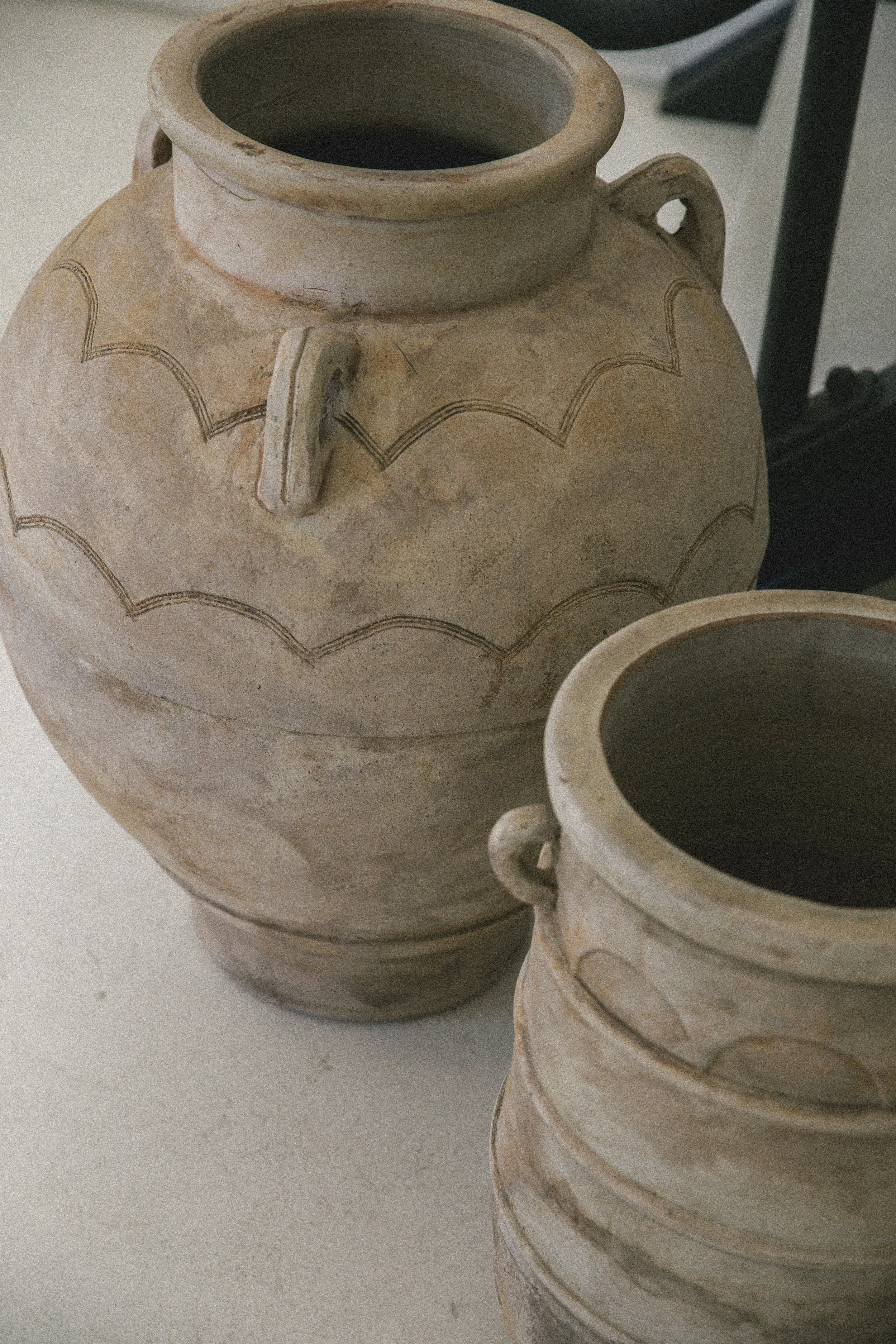 Two large beige pottery vases with decorative black markings and small handles, placed on a light-colored surface.