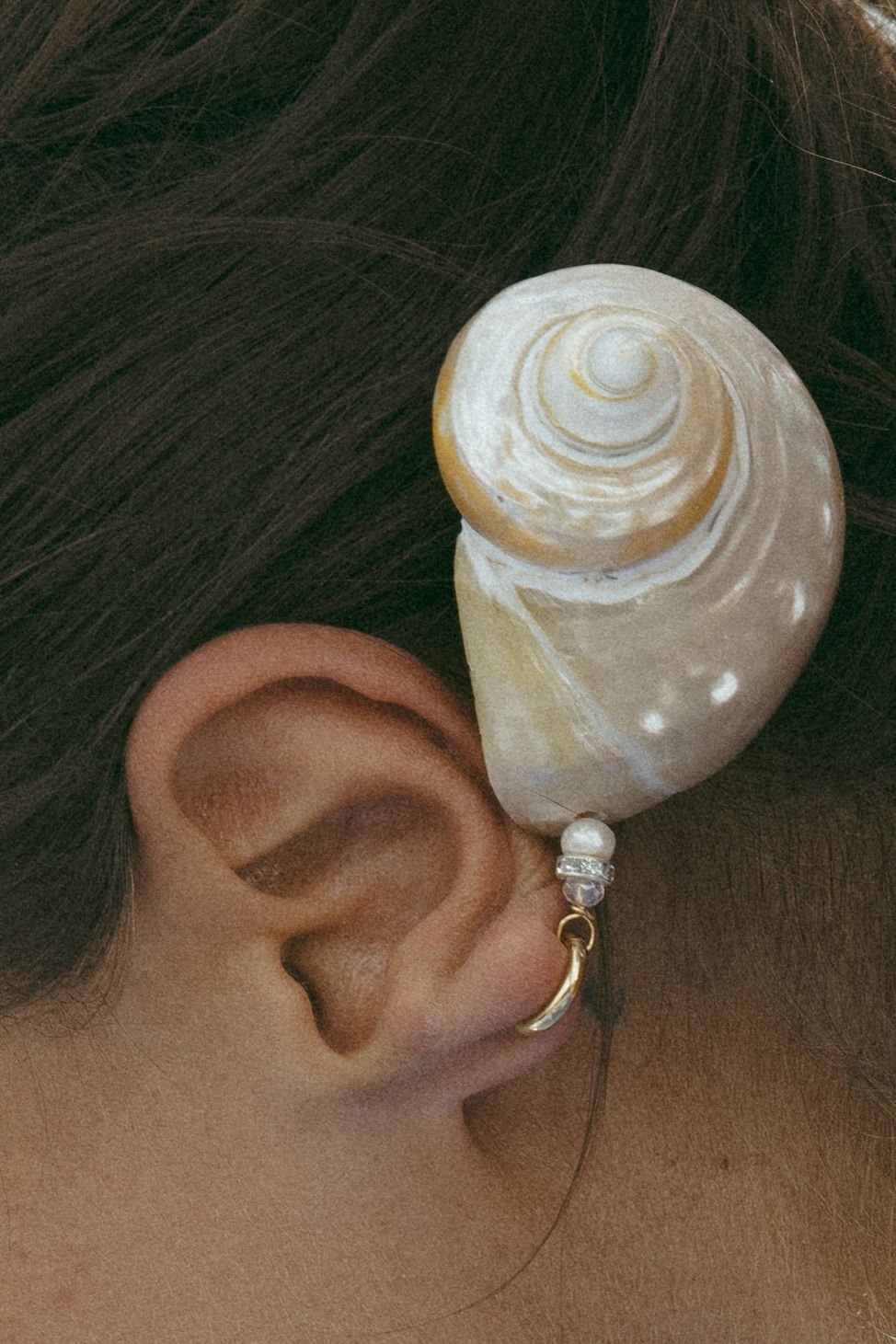 Close-up of a woman's ear with a decorated ear cuff featuring a shell attached to a black string with beads, including a pearl and a purple gemstone, above a dark hair background.