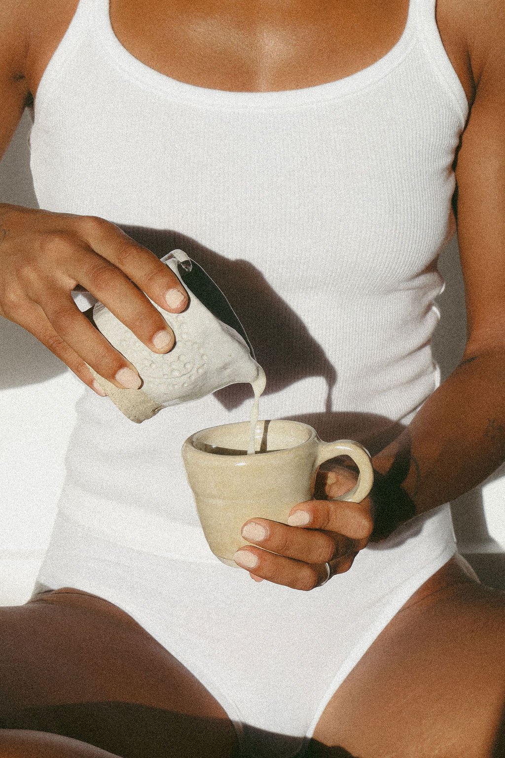 A person wearing a white tank top pours milk from a ceramic jug into a beige mug.