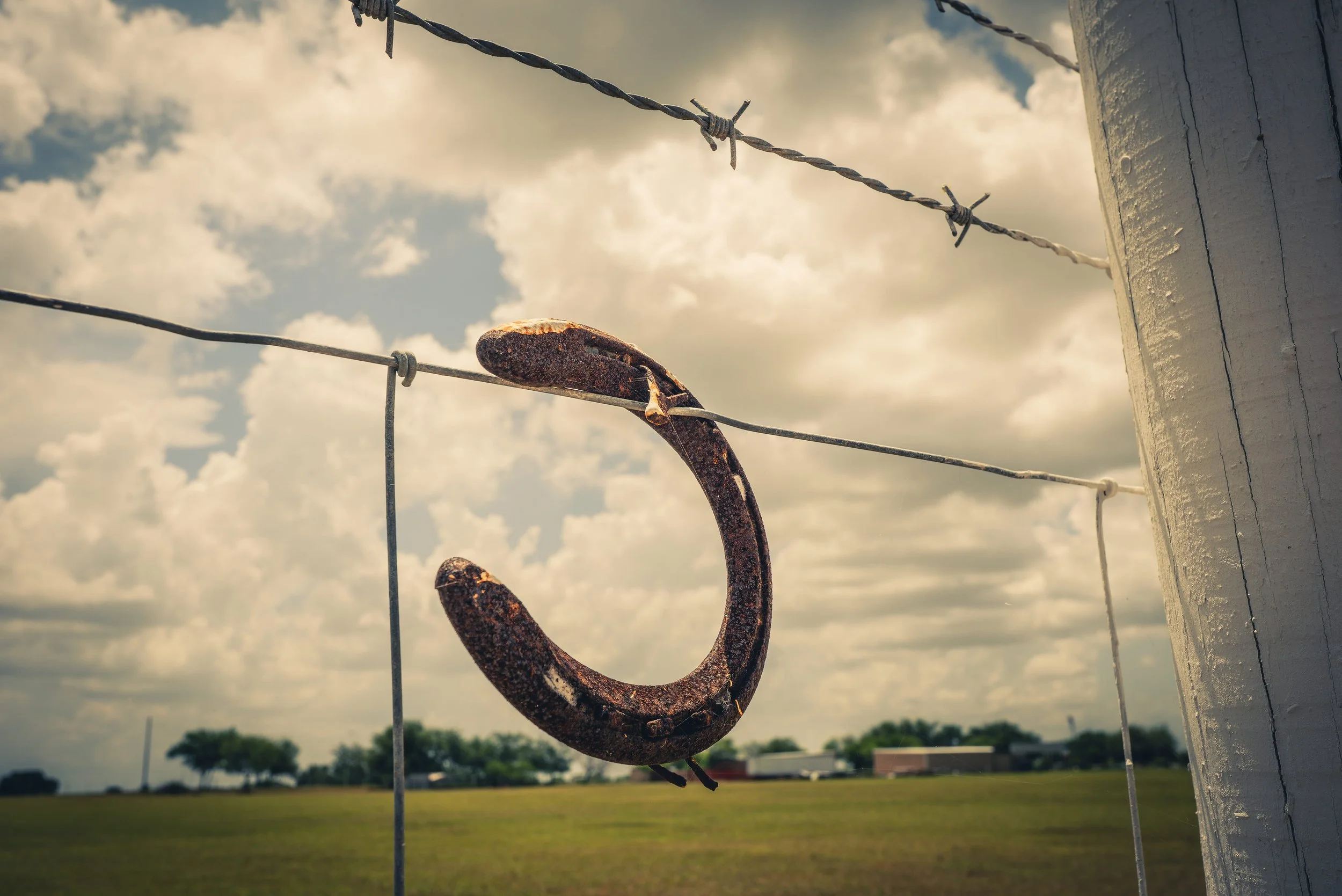 Close-up of rusty, weathered metal hook hanging on a wire fence, with a cloudy sky and open field in the background.
