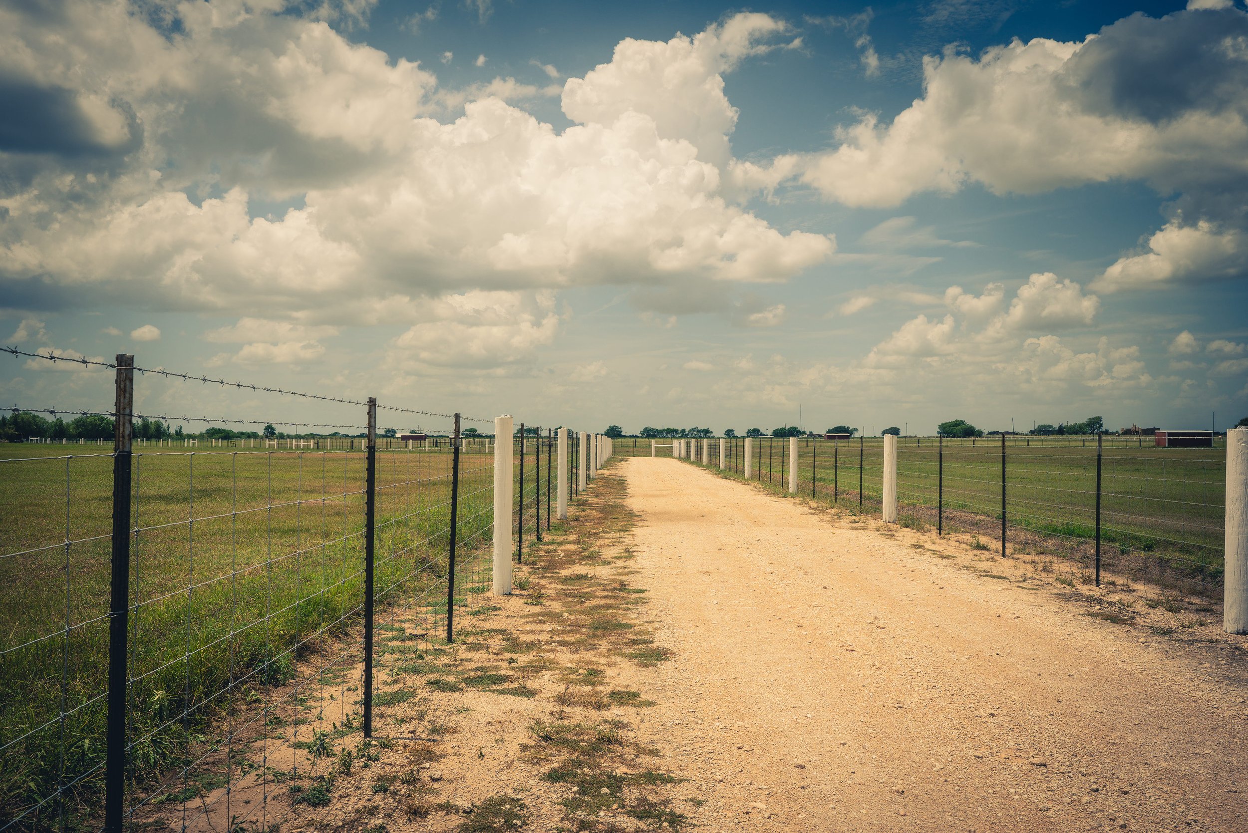 A dirt road runs through a grassy field bordered by fences on both sides, under a partly cloudy sky with scattered clouds.