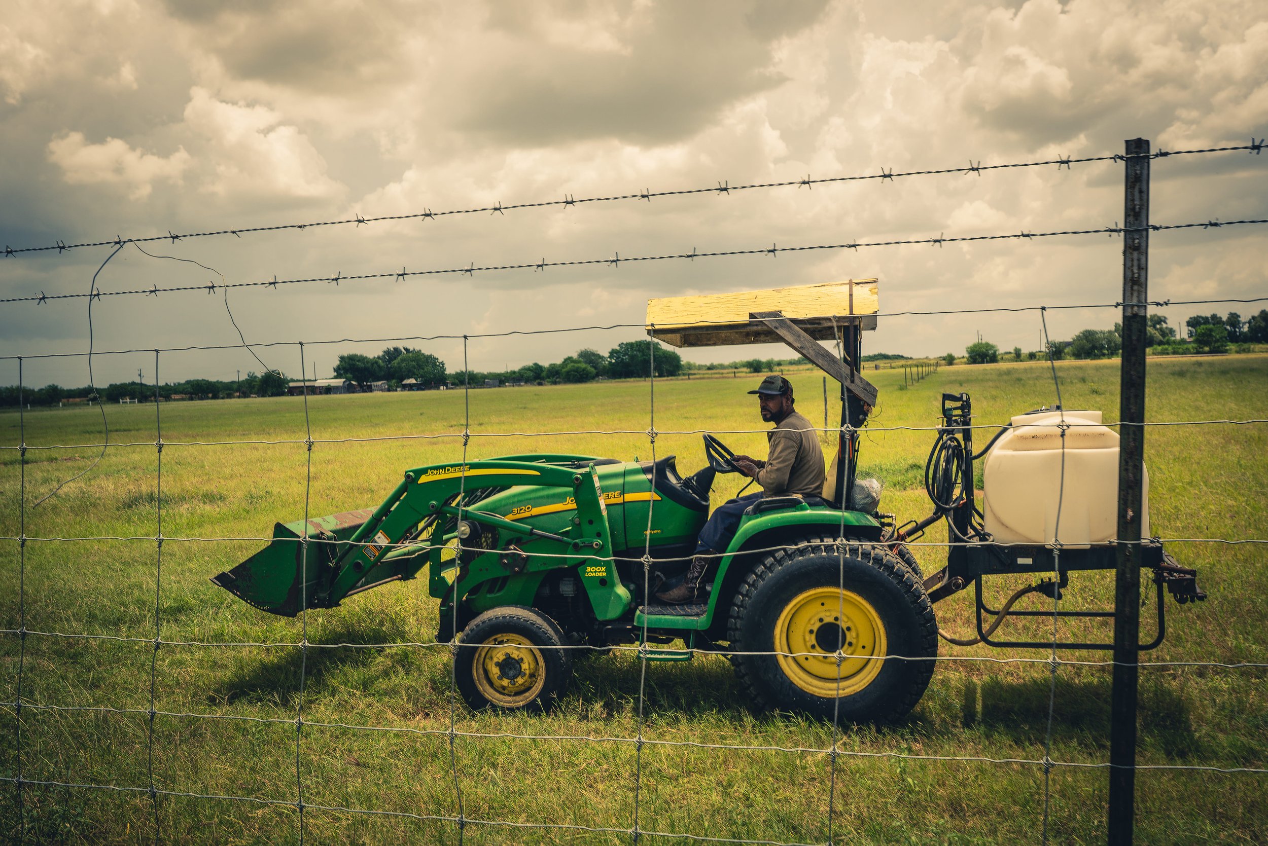 A man sits on a green John Deere tractor in a field, with a white tank attached, behind a barbed wire fence on a cloudy day.
