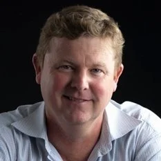 A man with curly blond hair wearing a light blue shirt, smiling against a dark background.