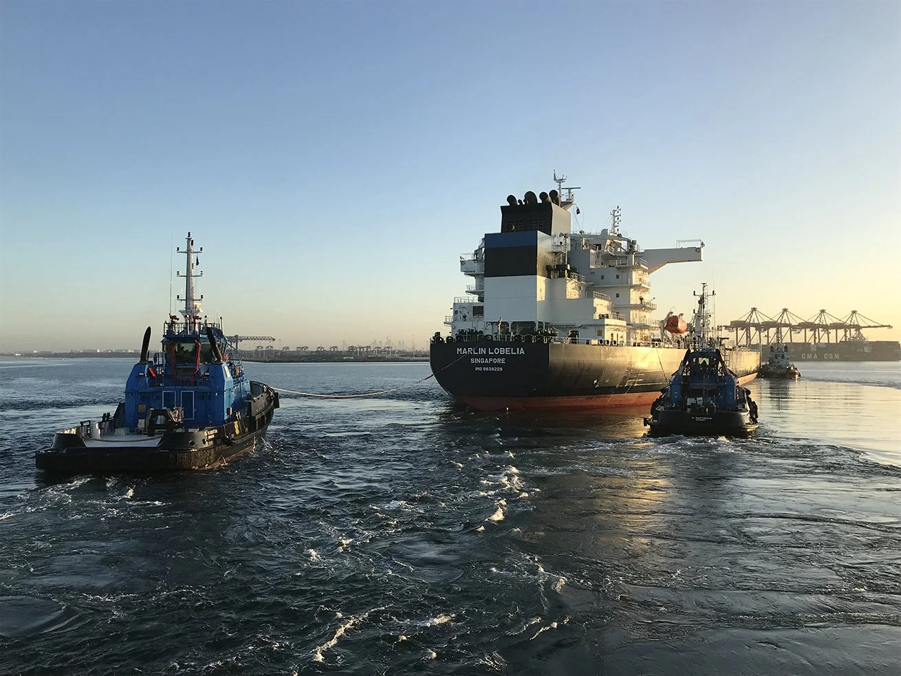 A large ship named Mari In Lobelia is being guided out of a port by two tugboats at sunset.