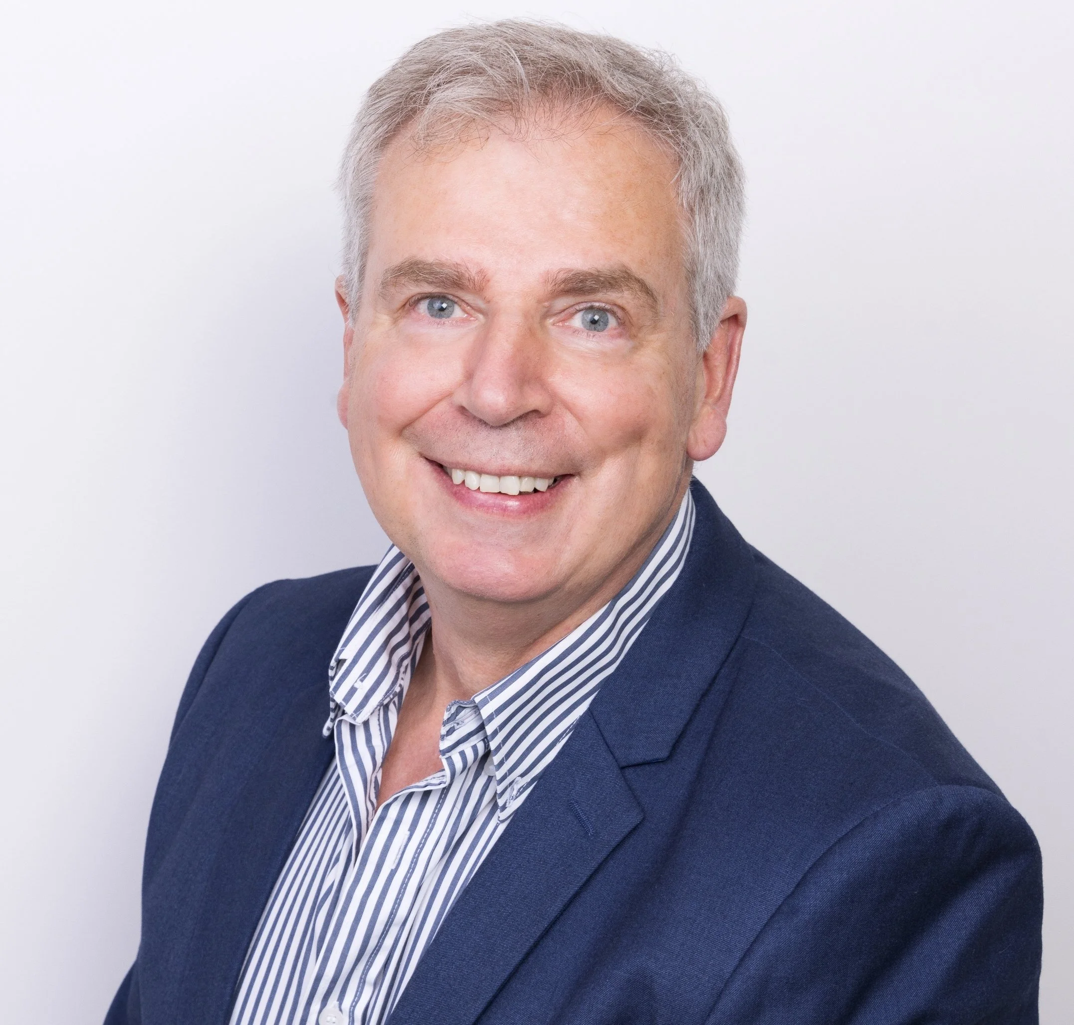 Close-up of a smiling man with gray hair, blue eyes, wearing a navy blazer and striped shirt against a white background.