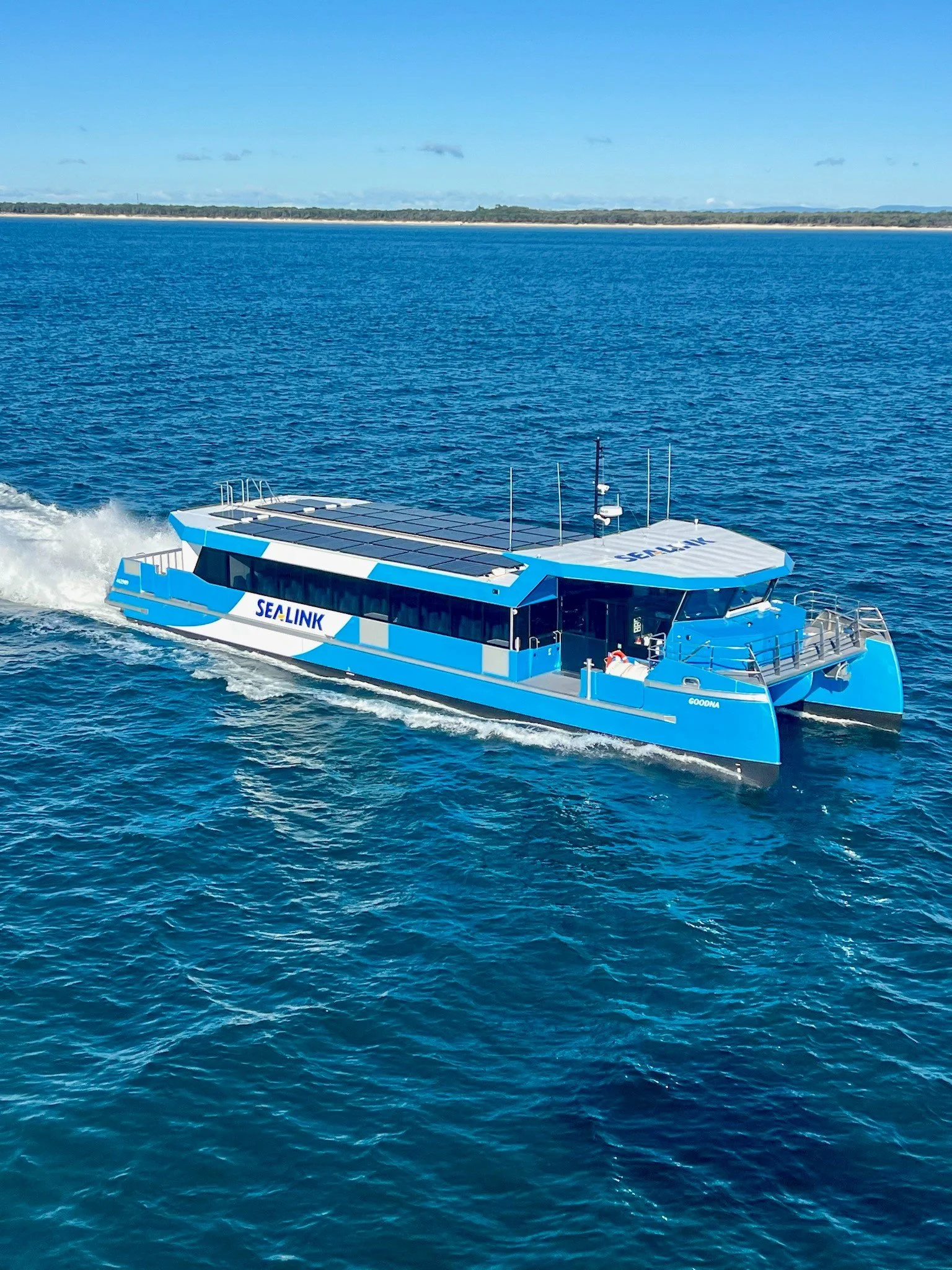A blue and white electric boat with solar panels on top, sailing on open water with a distant shoreline and blue sky in the background.