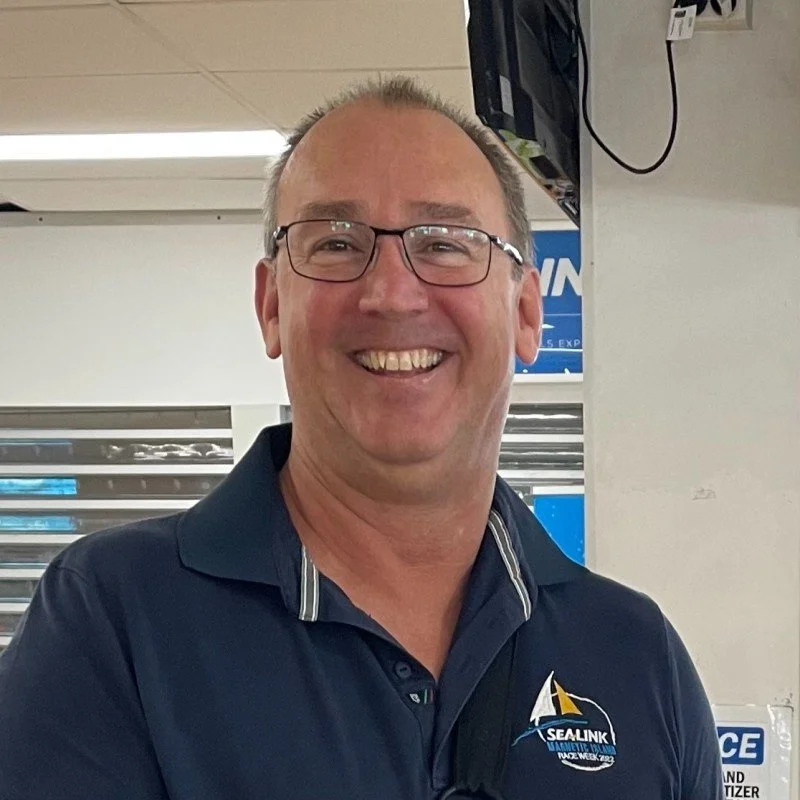 A happy middle-aged man with glasses, wearing a navy blue polo shirt with a Sealink logo, standing indoors with blinds and a wall-mounted electronic device in the background.