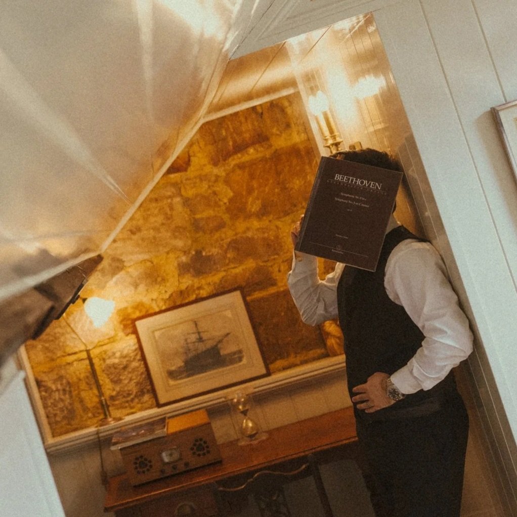 Person dressed in a white shirt and dark vest, holding a Beethoven vinyl record, standing in a cozy, warmly lit interior with wood-paneled walls, a framed painting of a ship, and a wooden sideboard with a vintage radio.