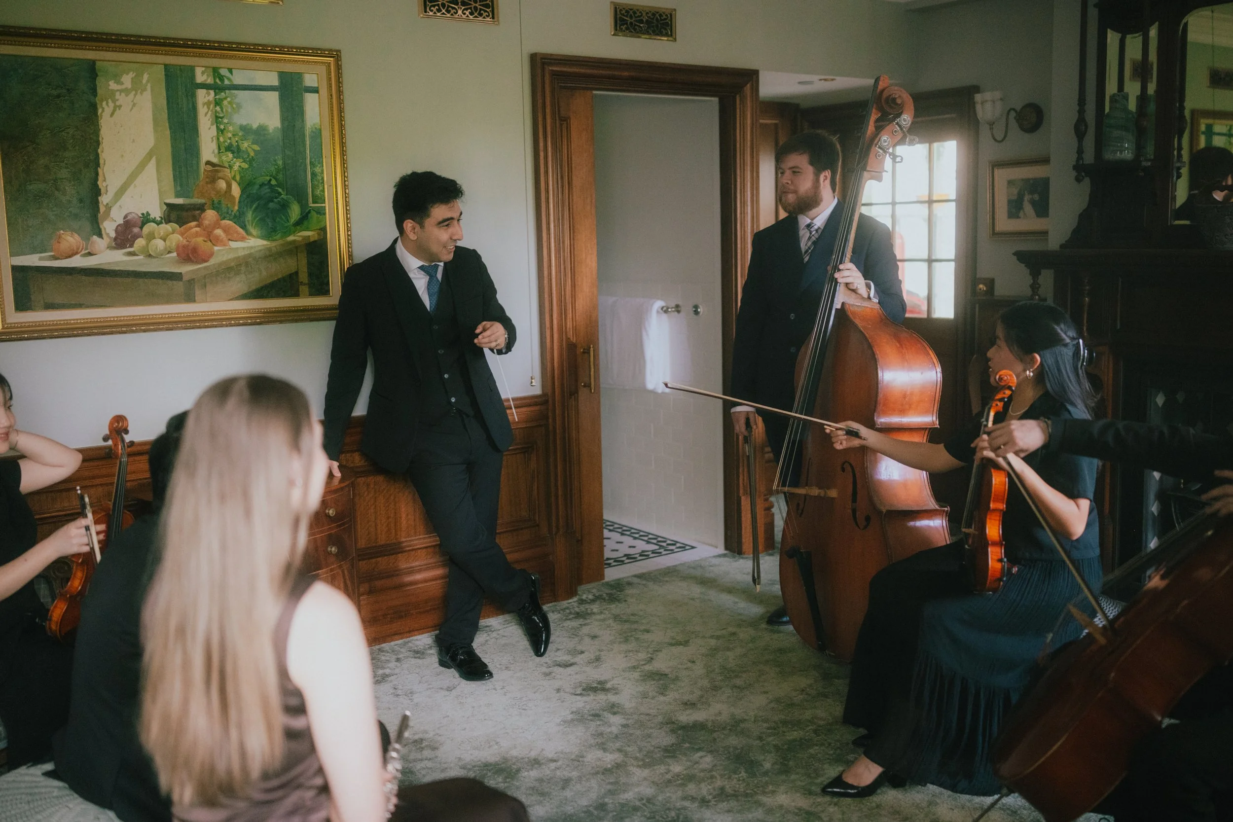 Two men in suits playing double bass and gesturing, as a woman with a violin listens in a living room with decorative paintings, wooden furniture, and a window in the background.