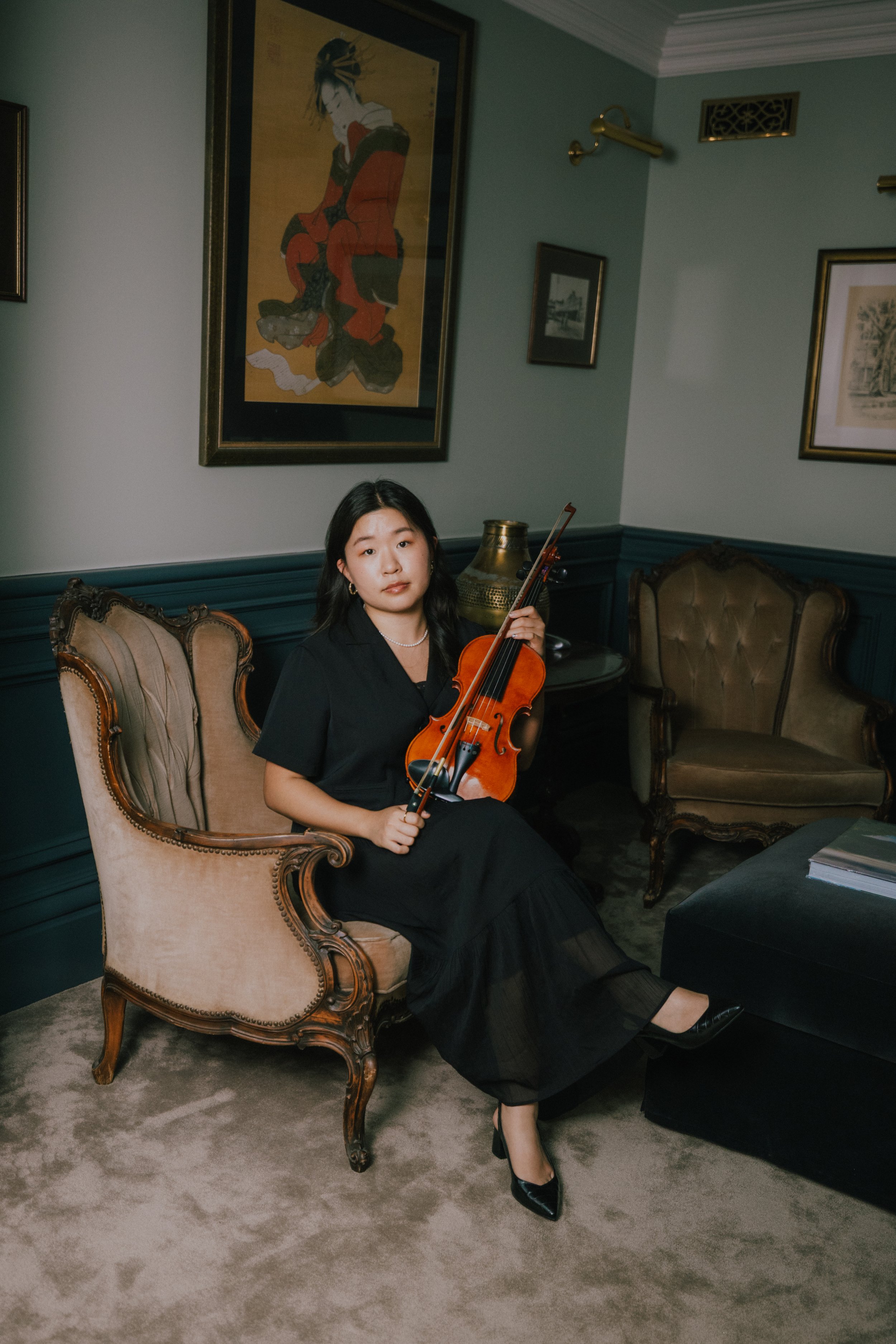 A woman in a black dress sitting on an ornate armchair in a room with classic furniture and framed artwork on the walls, holding a violin and bow.