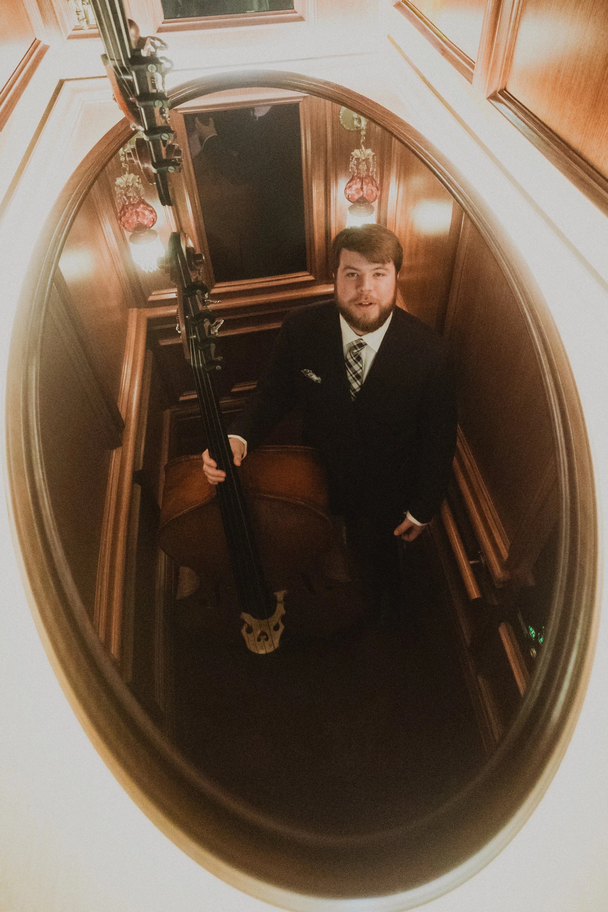 A man in a suit holding a double bass inside a wooden stairwell, seen through an oval-shaped mirror mounted on the ceiling.