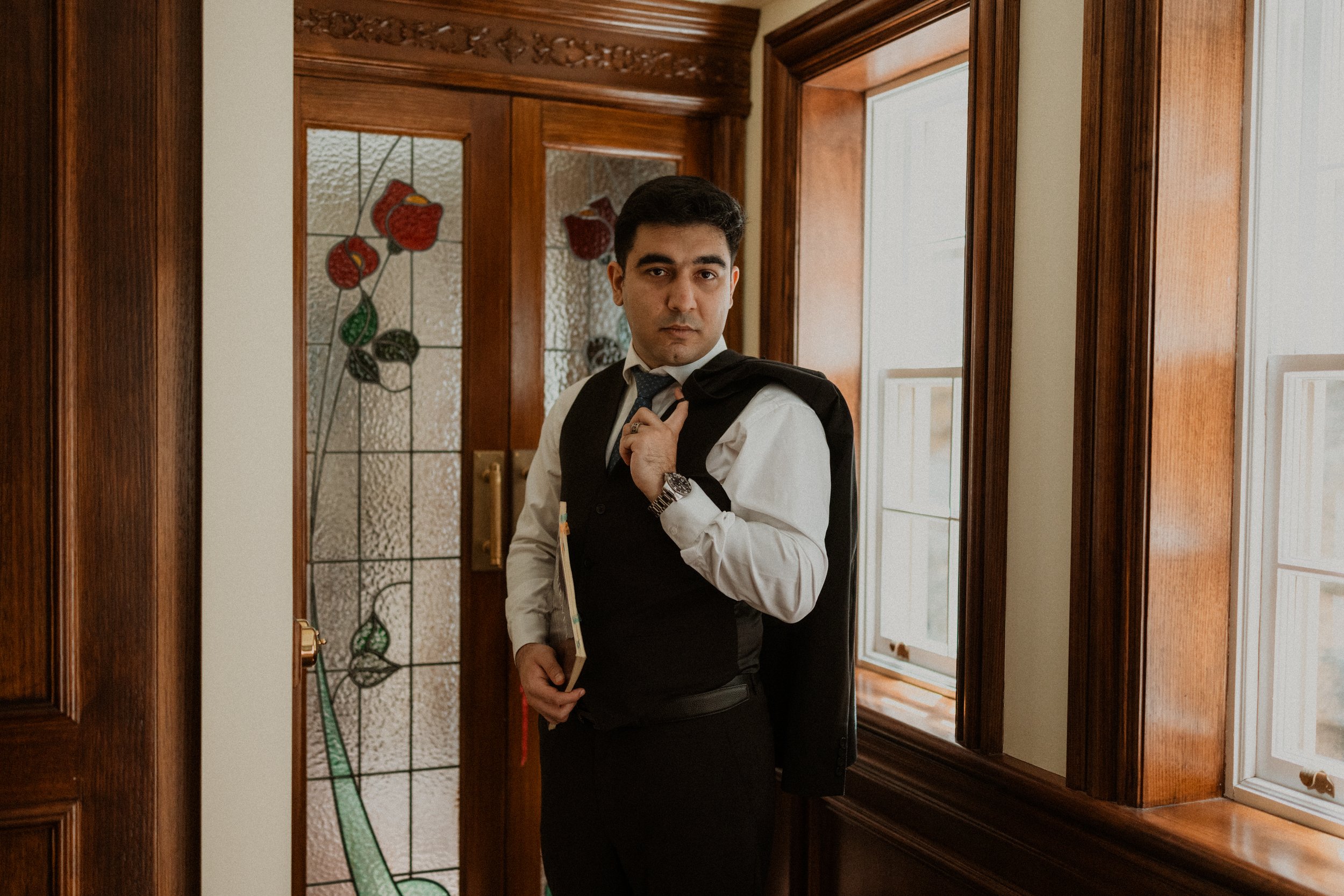 A man dressed in a white shirt and vest stands in a doorway, holding a jacket over his shoulder and a notebook, looking at the camera.