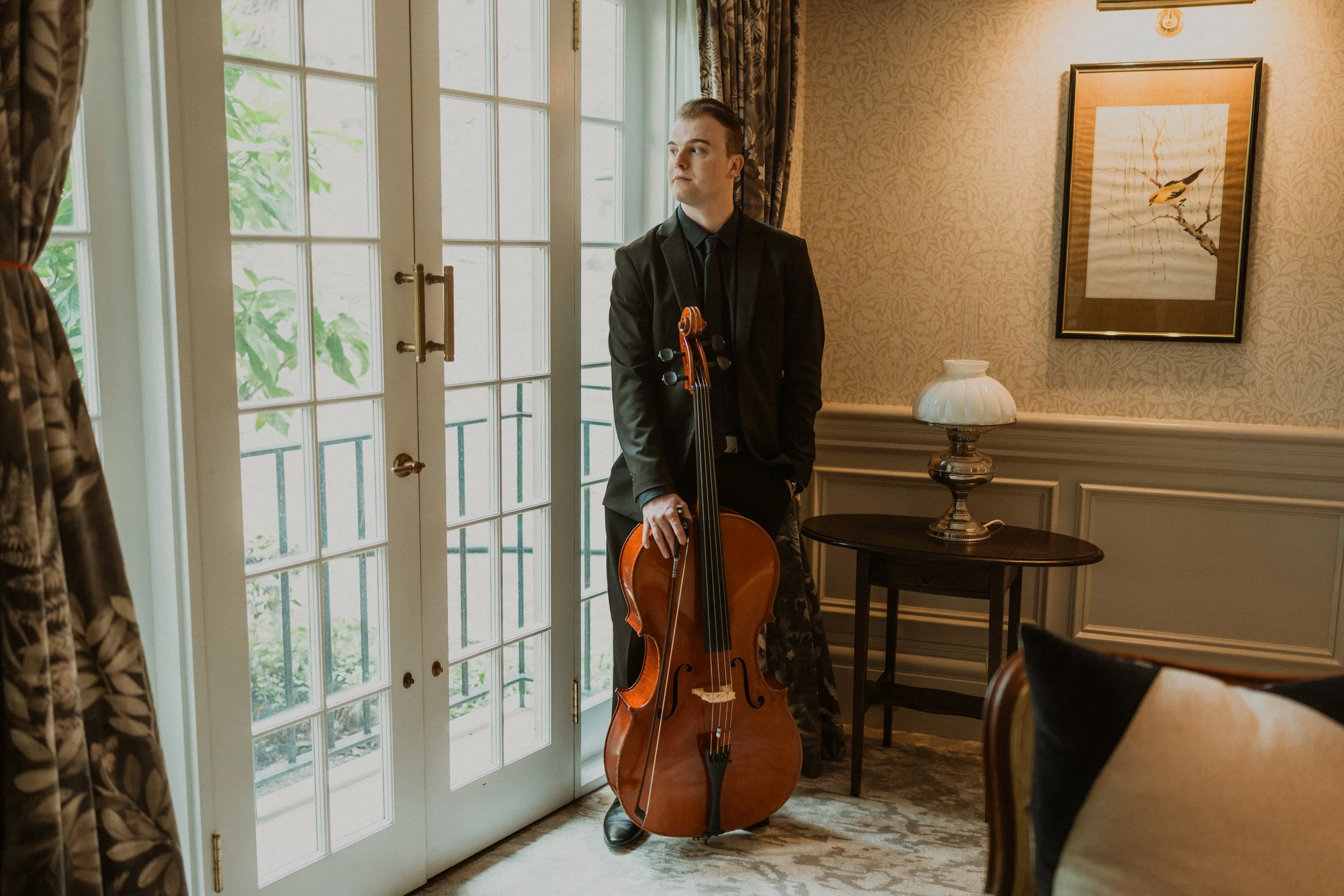 A young man in a black suit stands indoors near a glass-paneled door holding a cello upright. The room features floral patterned wallpaper, a framed artwork of a bird on a branch, a white decorative lamp on a small black table, and heavy patterned curtains.