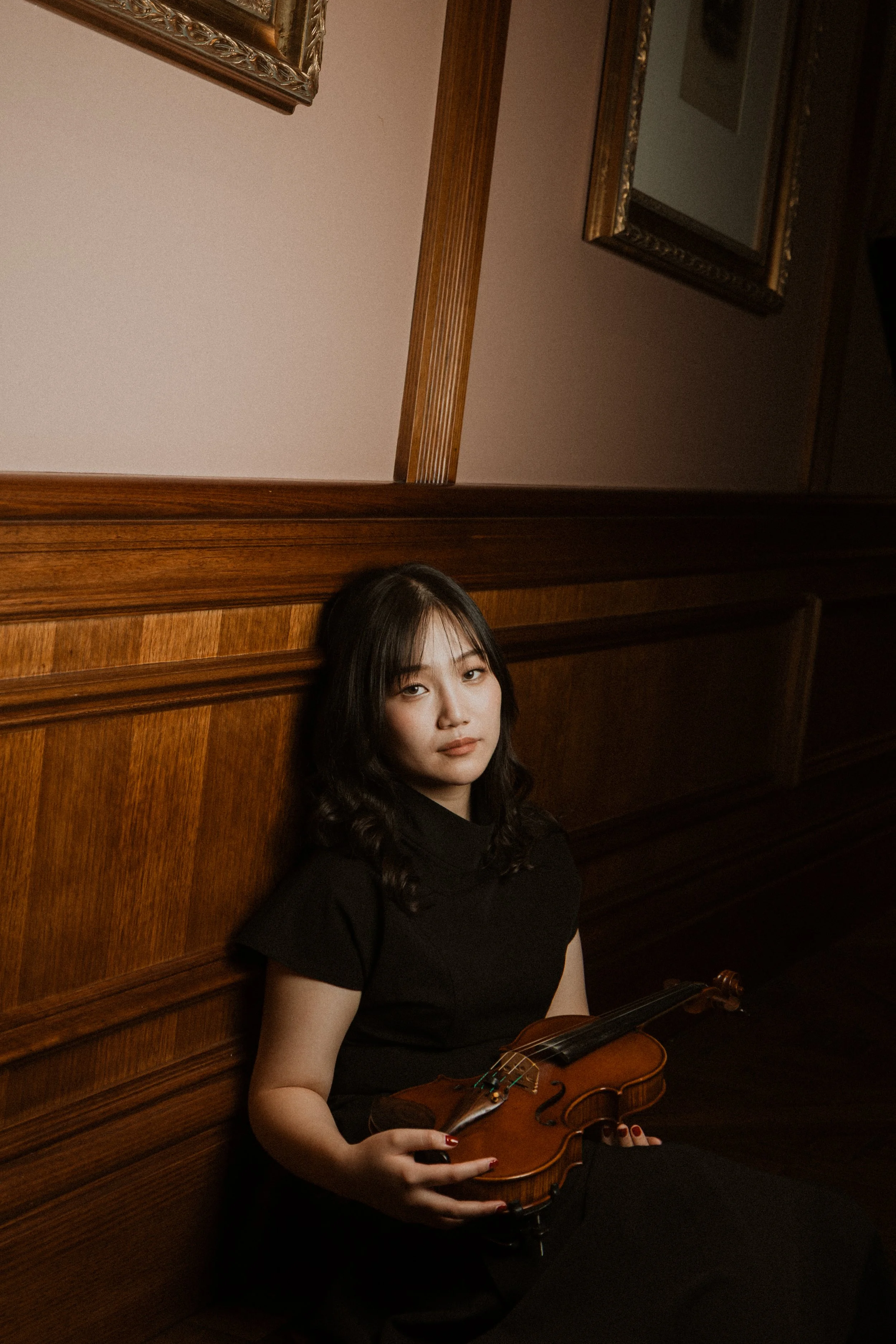A woman with dark hair and a black shirt sitting against a wooden-paneled wall, holding a violin in her lap, with the neck of the violin resting on her left hand.