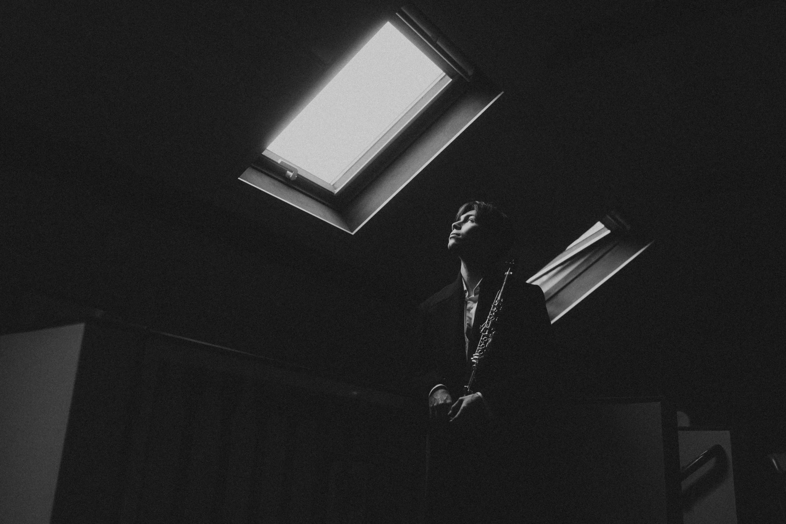 A person holding a clarinet looking up towards a skylight in a dark room.