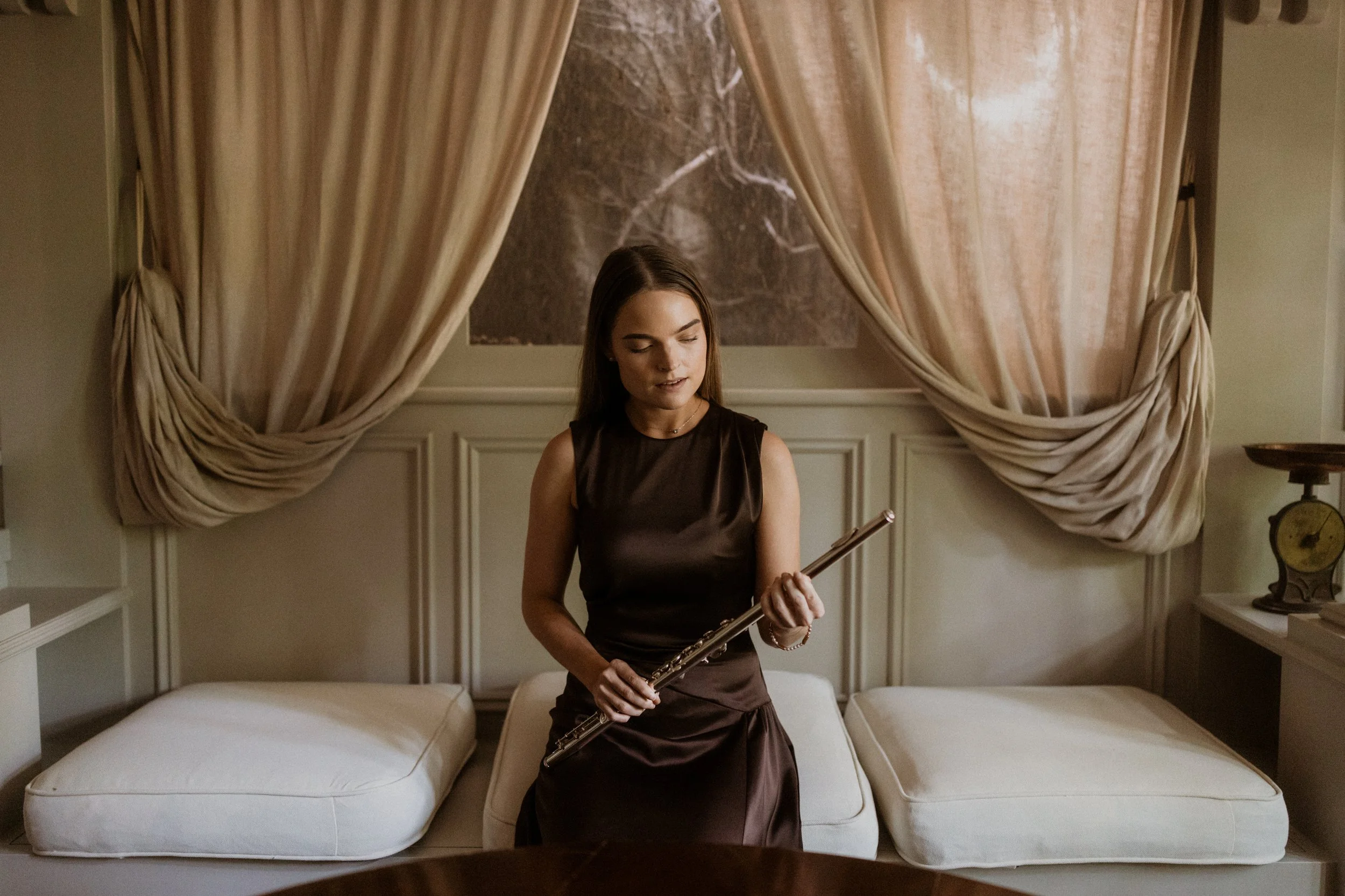 A woman with brown hair in a black dress stands in a bedroom, holding a flute, with bed and beige curtains behind her.