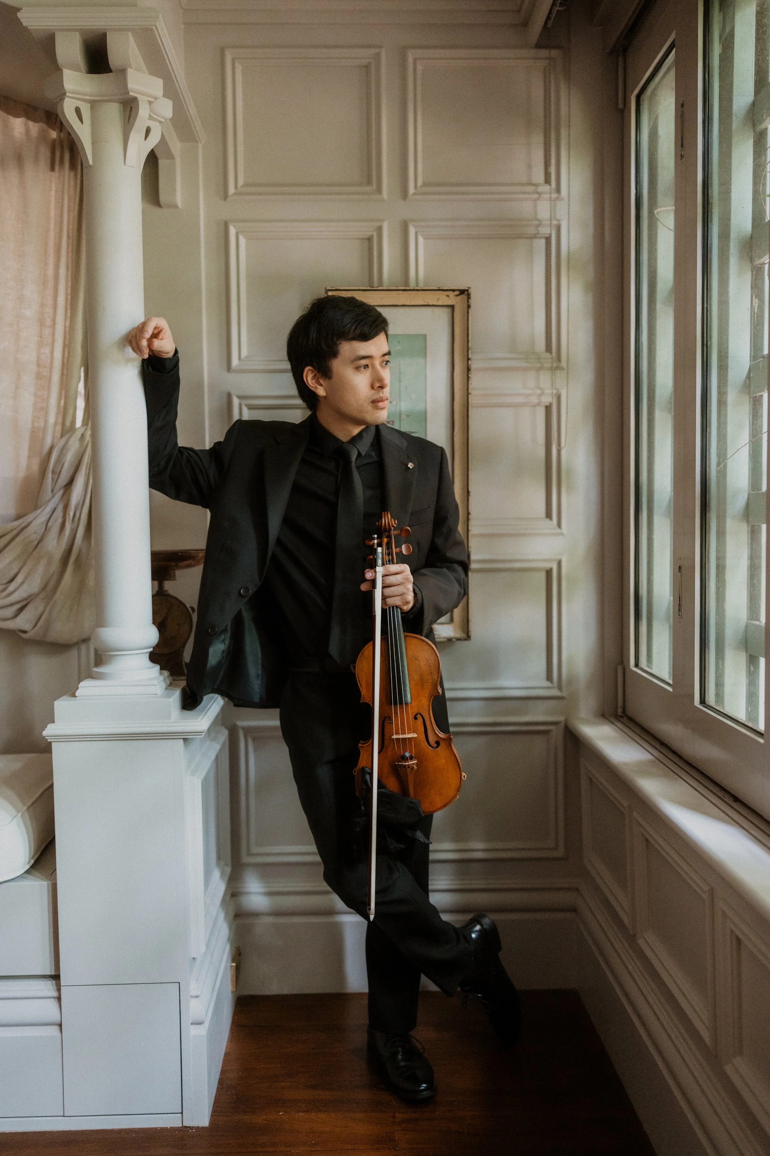 A man in a black suit holding a violin, standing in a room with white paneled walls and large windows, leaning against a white column with one arm and resting one foot on the wall.