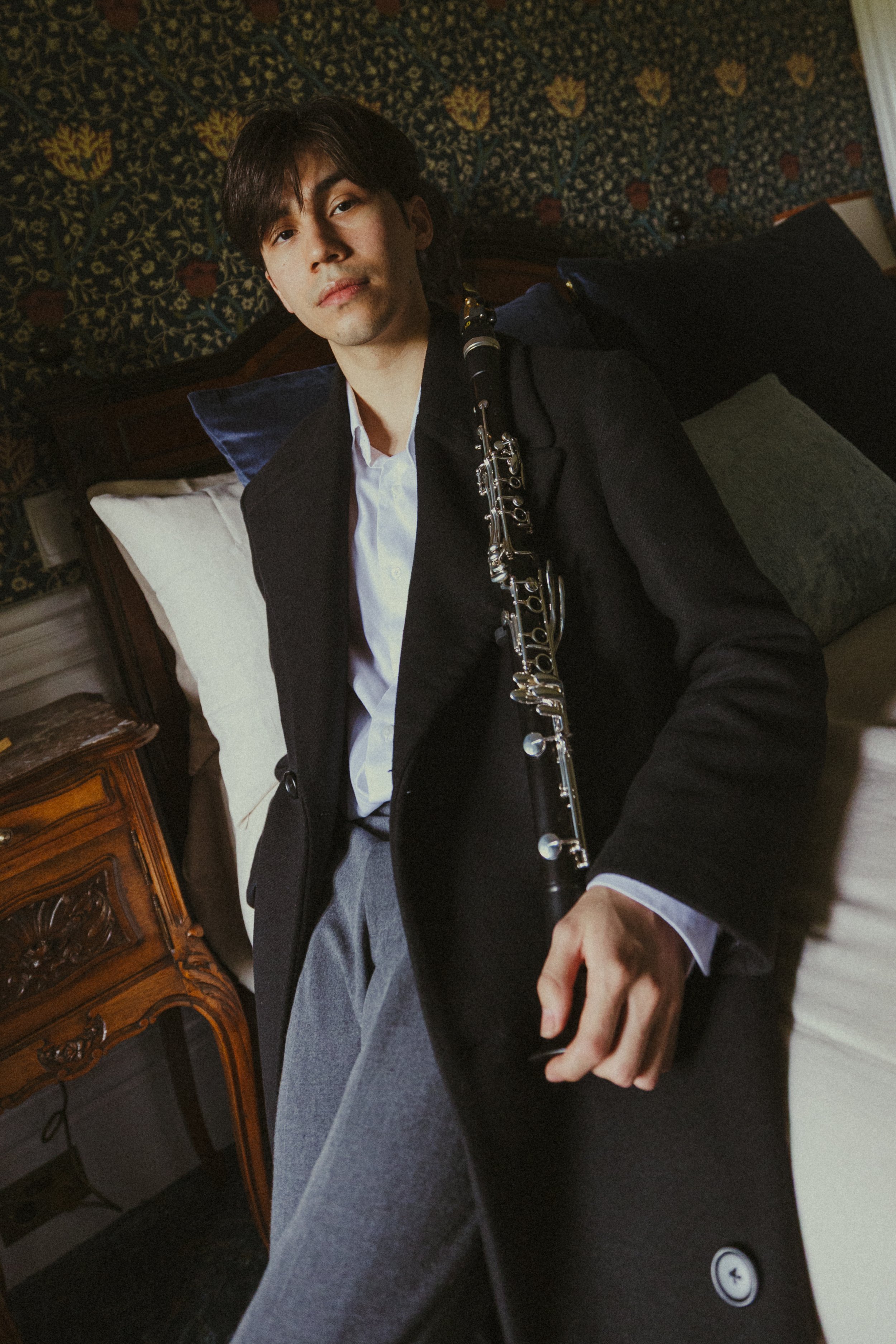 A young man with dark hair and fair skin lying in bed, wearing a black blazer and white shirt, holding a clarinet, with a green pillow and a patterned tapestry behind him.