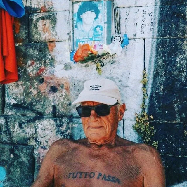An elderly man wearing sunglasses and a white cap, with tattoos on his chest, sitting shirtless outdoors against a stone wall with a photo of a young person, flowers, and some handwritten notes above his head.