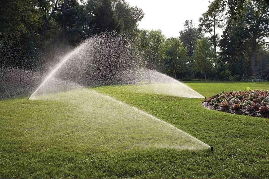 Sprinklers watering a lush green lawn in a park or garden with trees in the background.