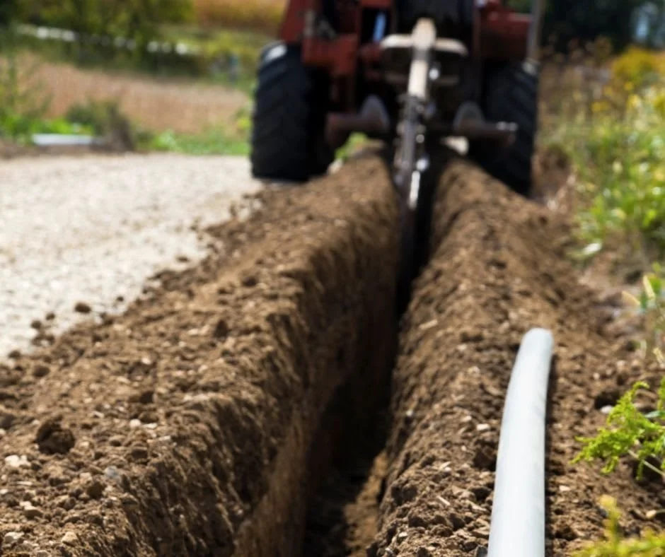 Close-up of a trench being dug in the ground with a small tractor in the background