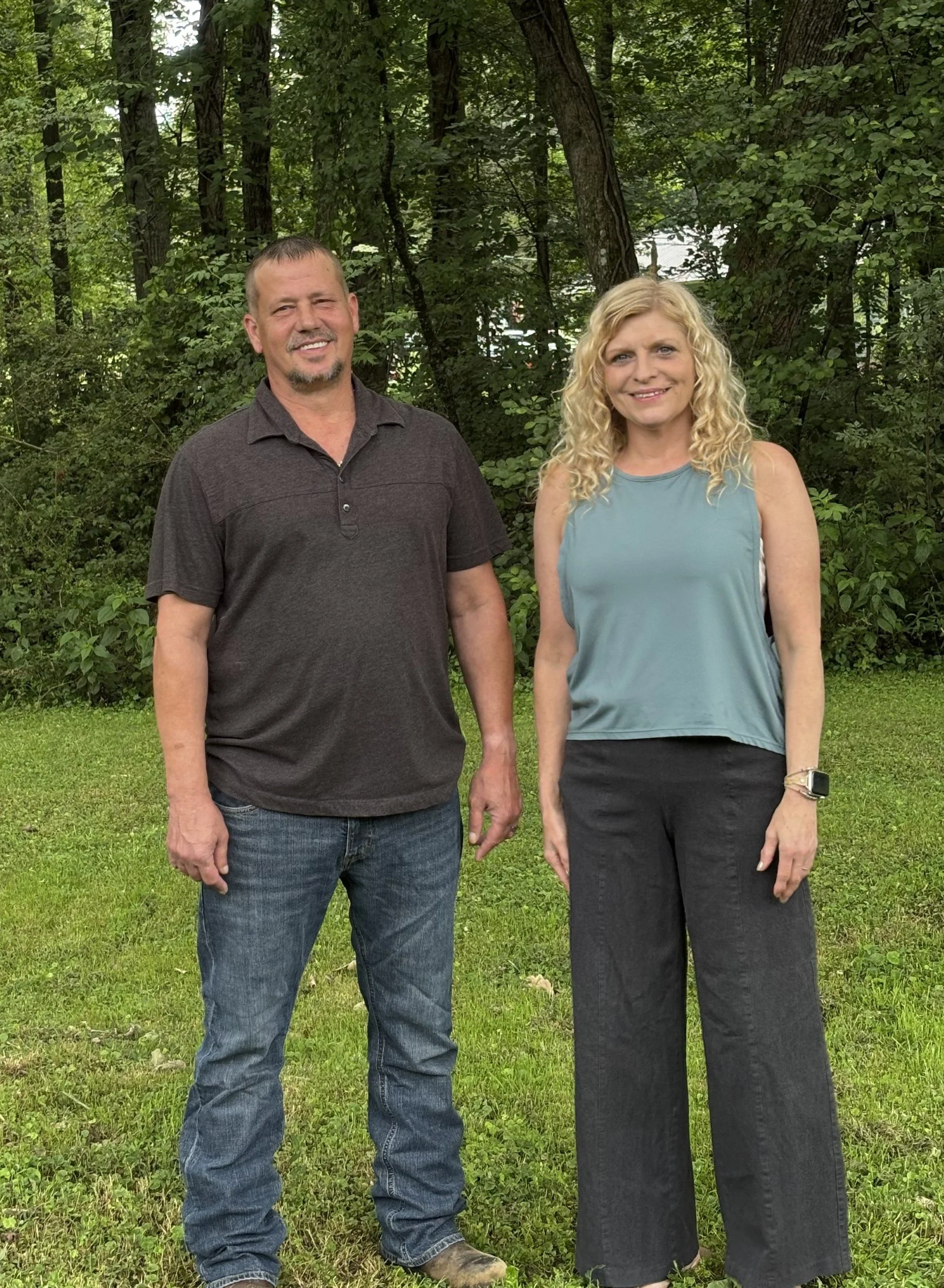 A man and a woman standing outdoors in a green, wooded area, holding hands and smiling at the camera.