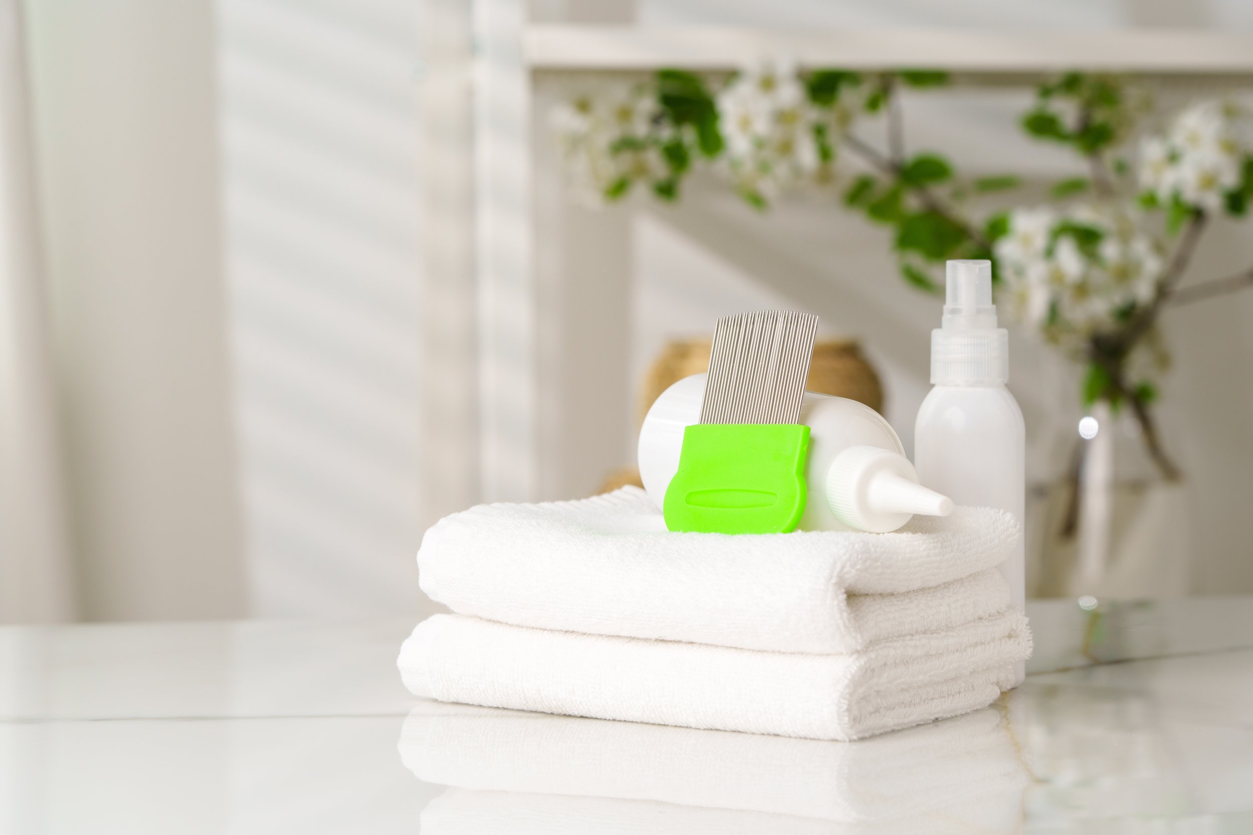A neatly folded white towel with various toiletries on top, including a spray bottle, a tube, a comb, and a small container, with a blurred background of green plants and white shelves.