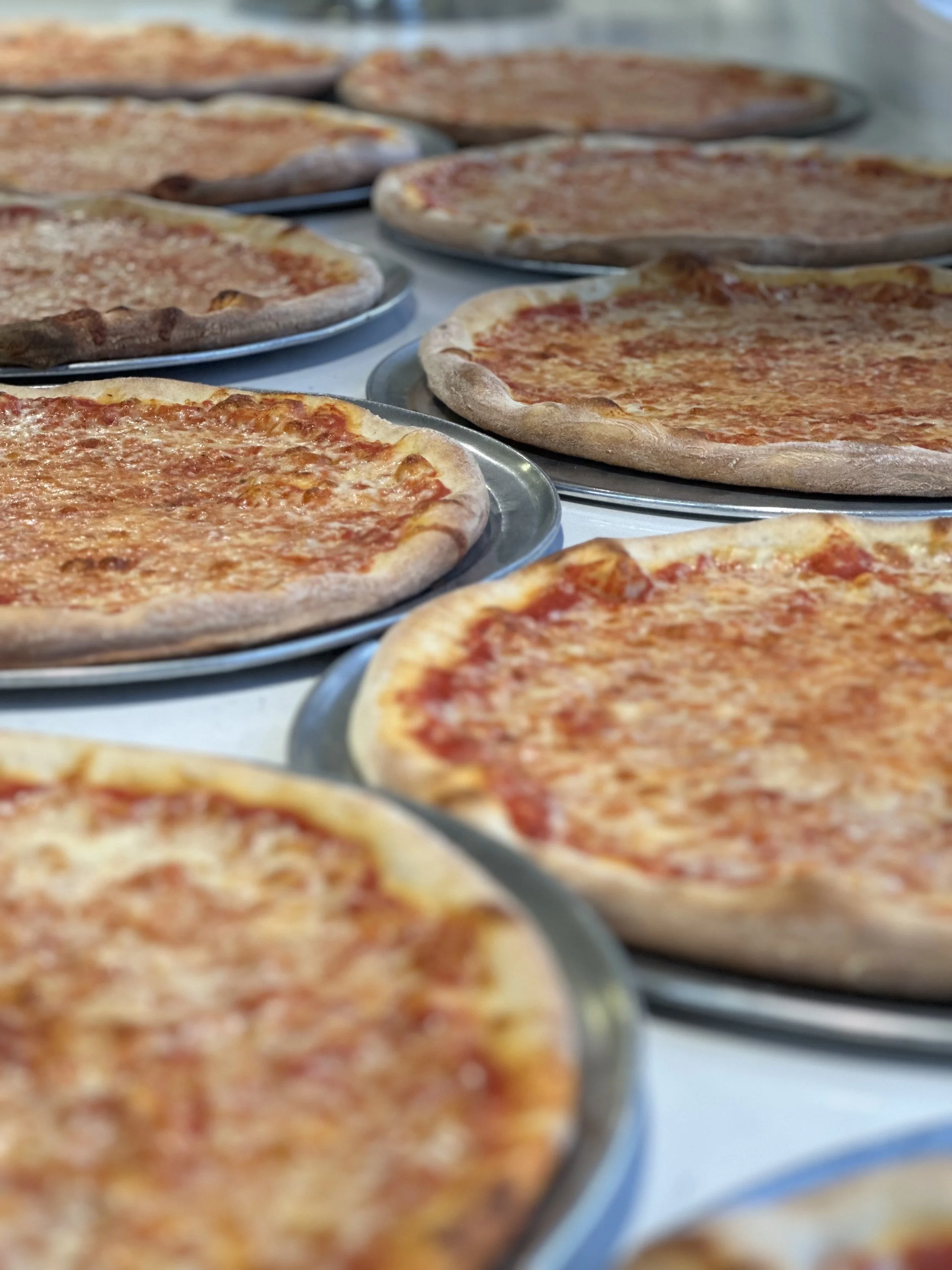 Close-up of multiple uncooked cheese pizzas on metal trays.
