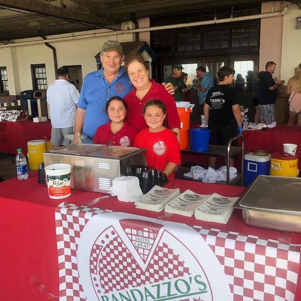 A family of five poses behind a food stand at an event, with two adults and three children smiling. The stand has a red checkered banner with a pizza logo and the name 'PANDAZZO'S'. There are food items, napkins, cups, and cold drink dispensers on the table, with a crowd of people in the background.