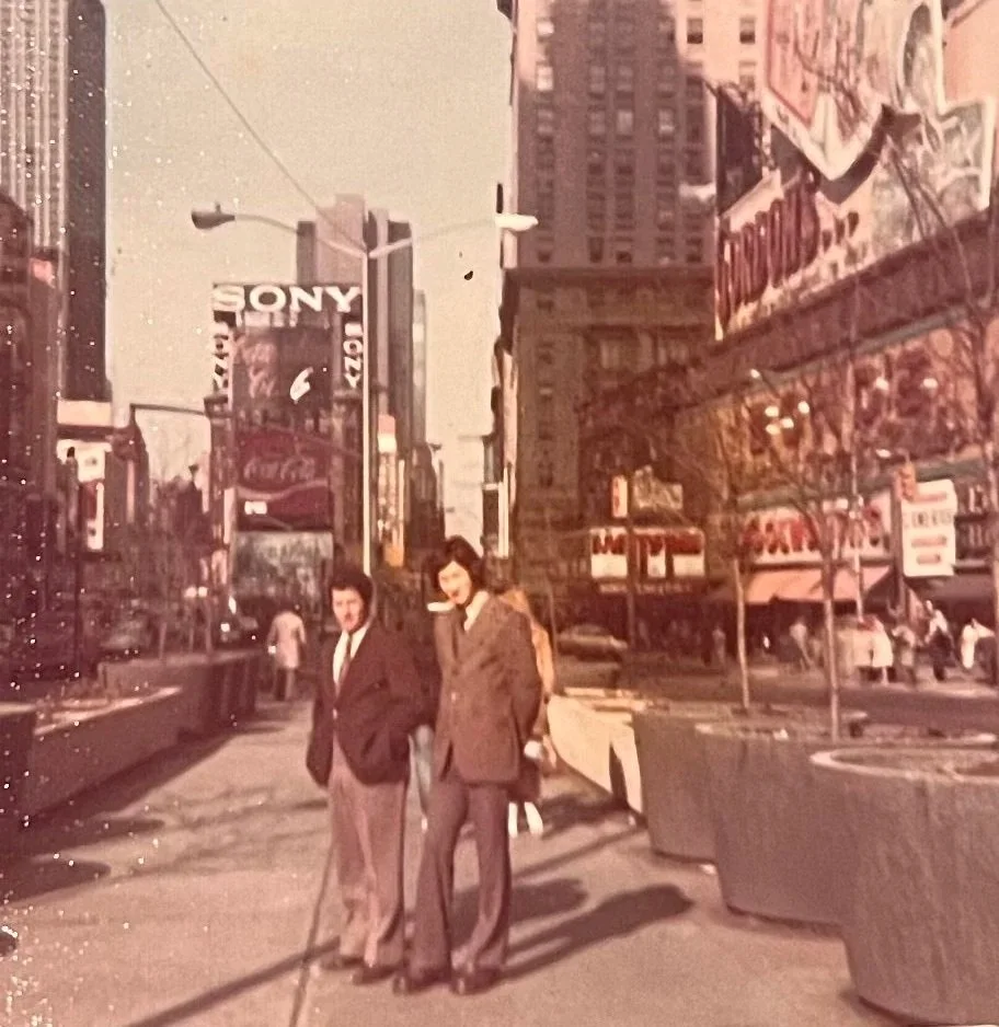 Two women standing on a busy city sidewalk with tall buildings, billboards, and a bus in the background.
