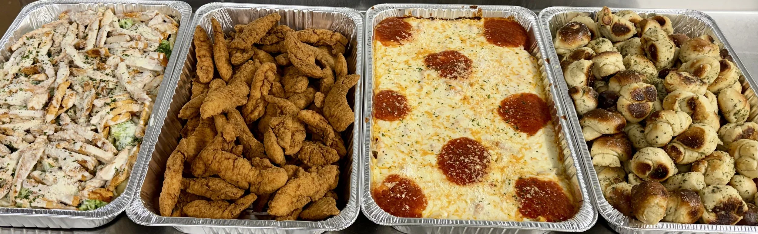 Four aluminum trays filled with different foods: a salad with shredded chicken, lettuce, and cheese; crispy fried chicken strips; a baked cheese and pepperoni casserole; and a tray of baked sausage rolls.