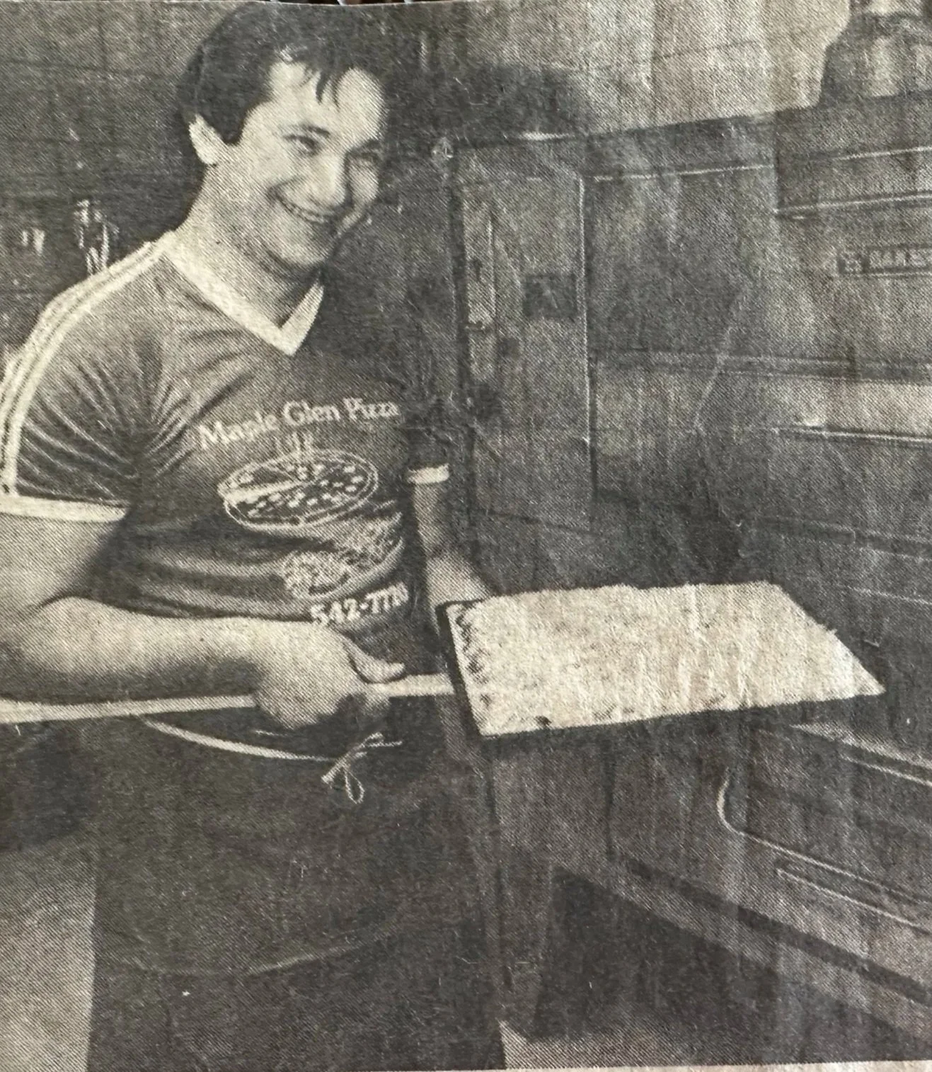 Black and white photo of a young man in a sports jersey smiling and sitting at a table with an open notebook, with a brick wall in the background.