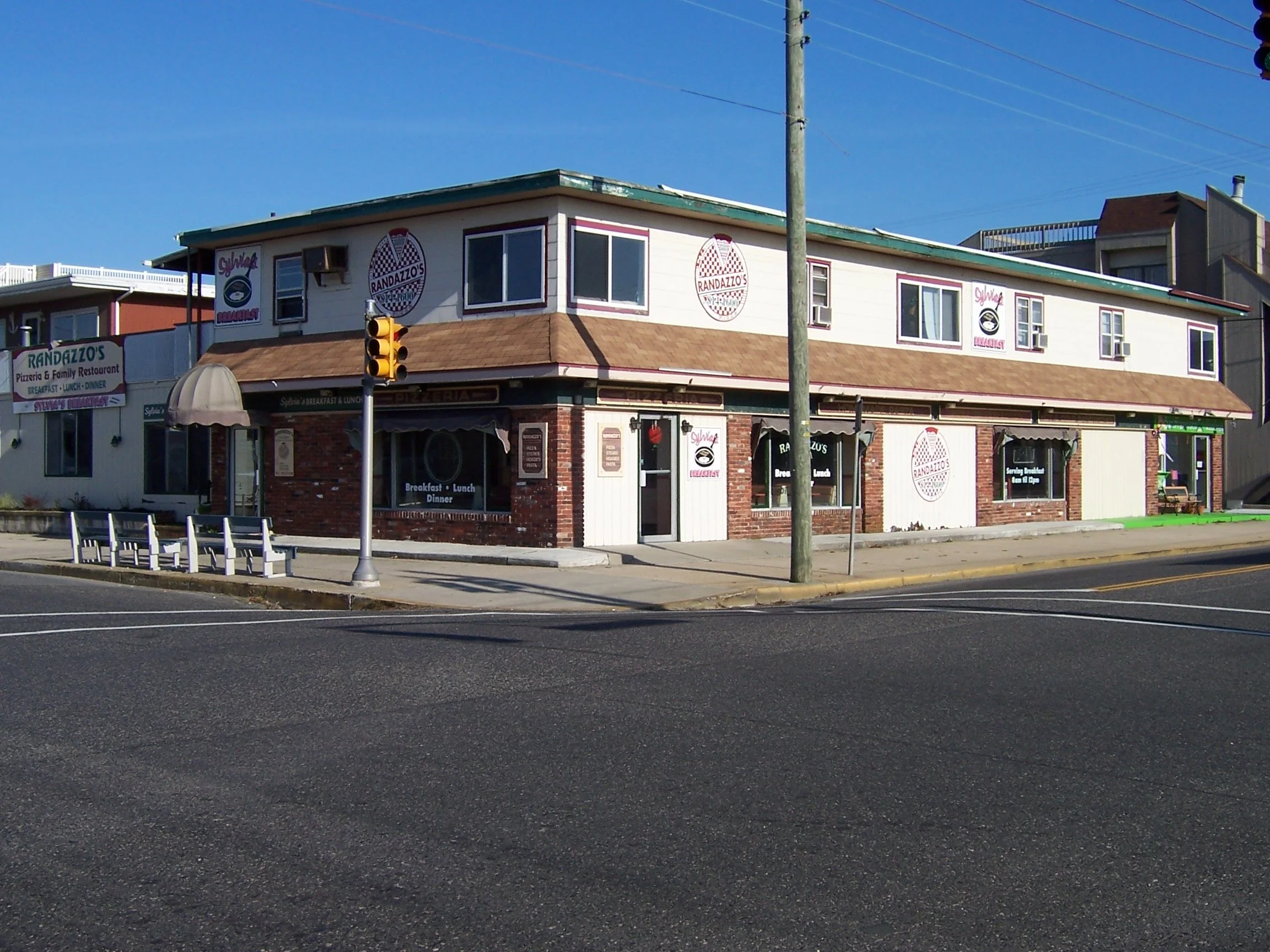 A two-story restaurant building with a sign reading 'Randazzo's Pizzeria & Family Restaurant' on the front. The building has white siding, brick accents, and several windows. There is a traffic light and a utility pole in the foreground, with a clear blue sky above.